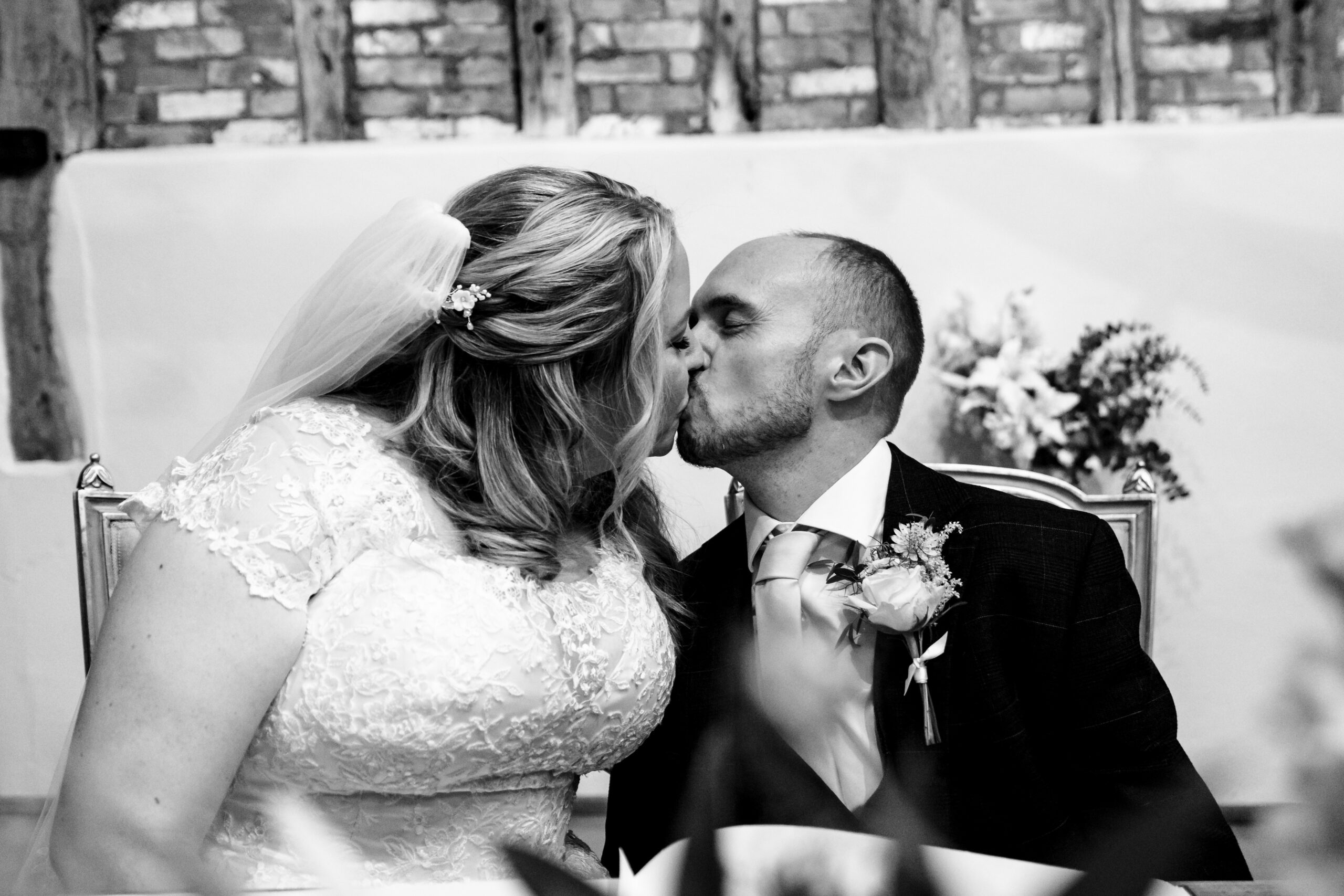 A black and white image of the bride and groom kissing. They are sat down about to sign their legal paperwork. 