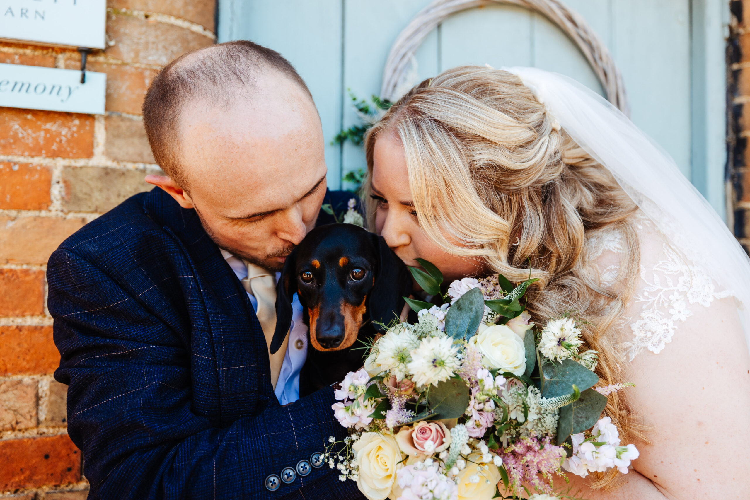 The bride and groom are huddled together with their dog in the middle. The dog is looking at the camera and the bride and groom are kissing the top of the dogs head.