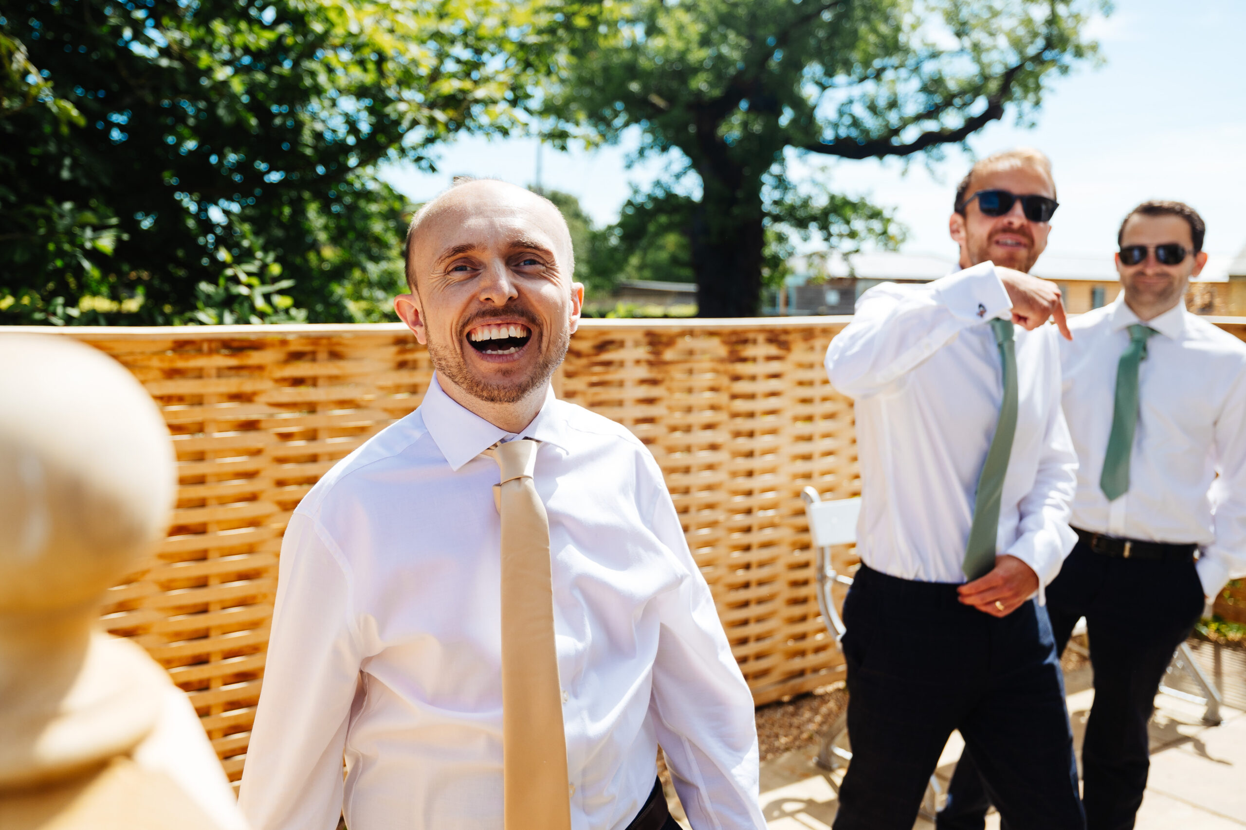 A man (the groom) outside with two of the groom's party. He is smiling and looking at the camera wearing a white shirt and yellow tie.