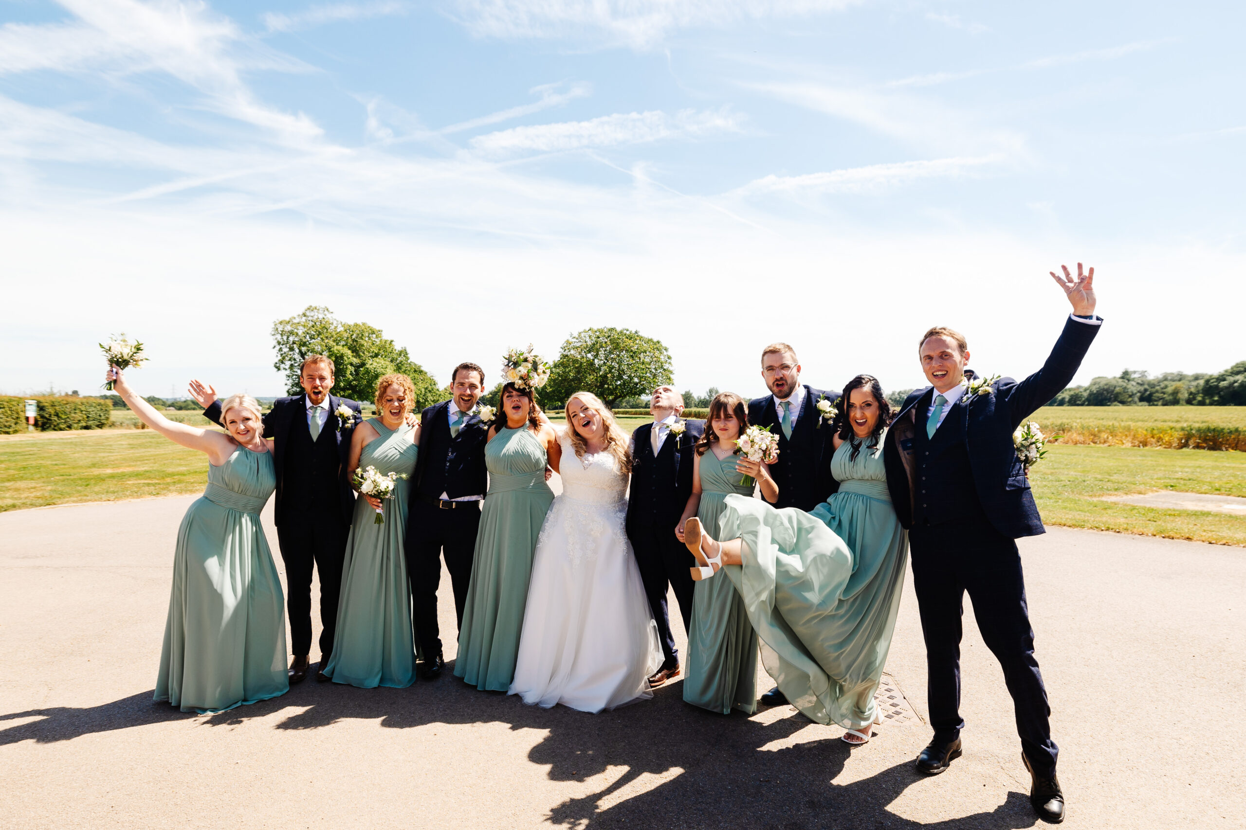 A group shot of the bridal party. They have their hands in the air and are smiling and look excited and like they are having a good time. They are outside and the sky is blue.