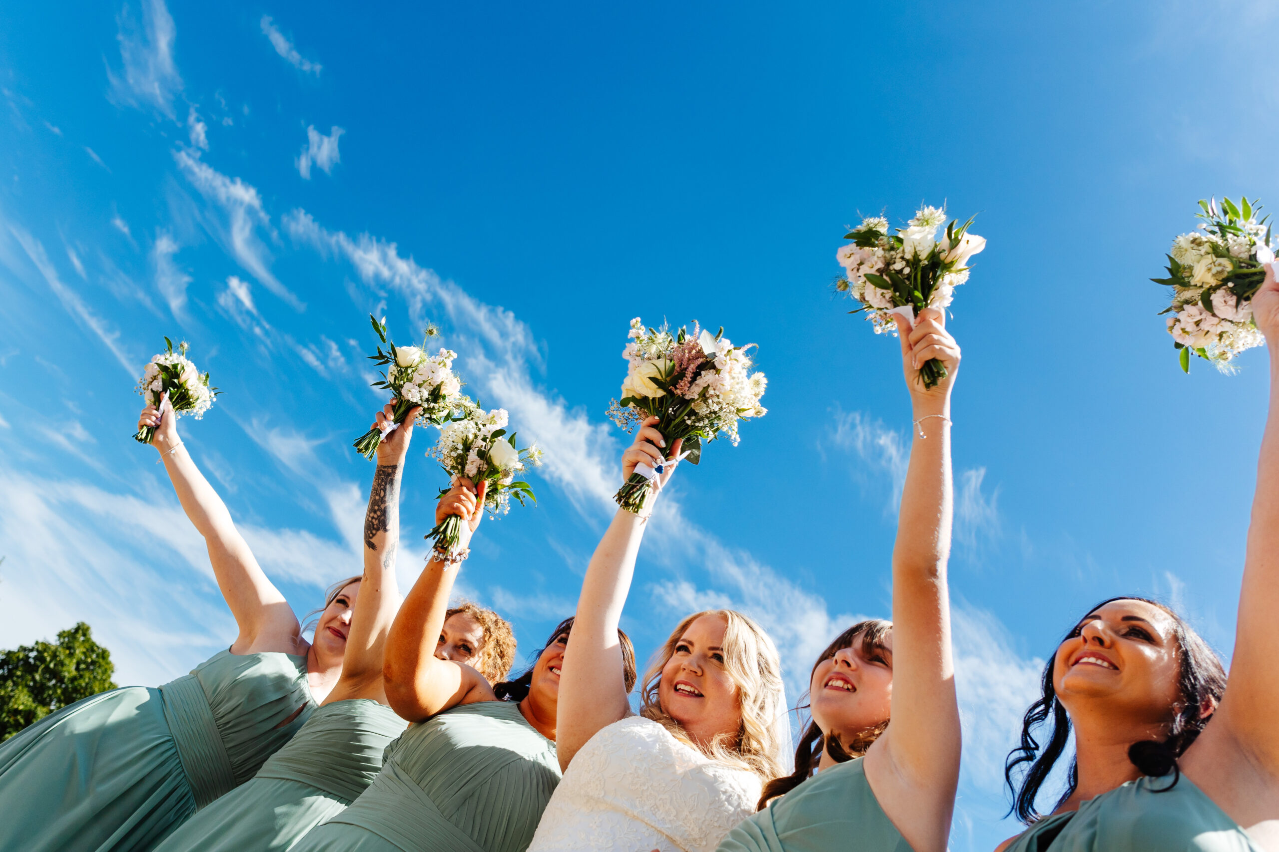 An image of the bridal party lifting up their bouquets to the sky. The sky is bright blue.