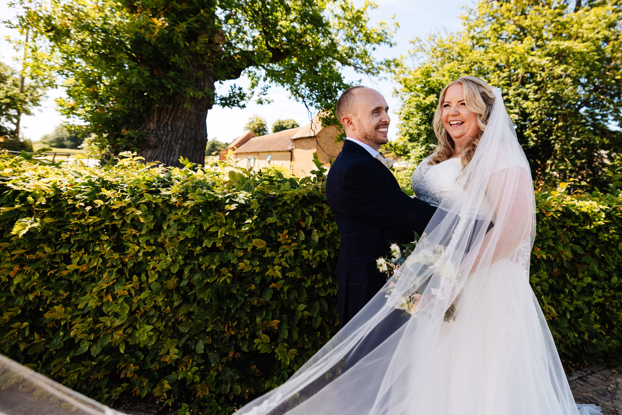 The bride and groom outside near some greenery of trees and a hedge. They are looking in the distance and smiling. The groom has his hands around the bride's waist and her veil is floating in the wind.