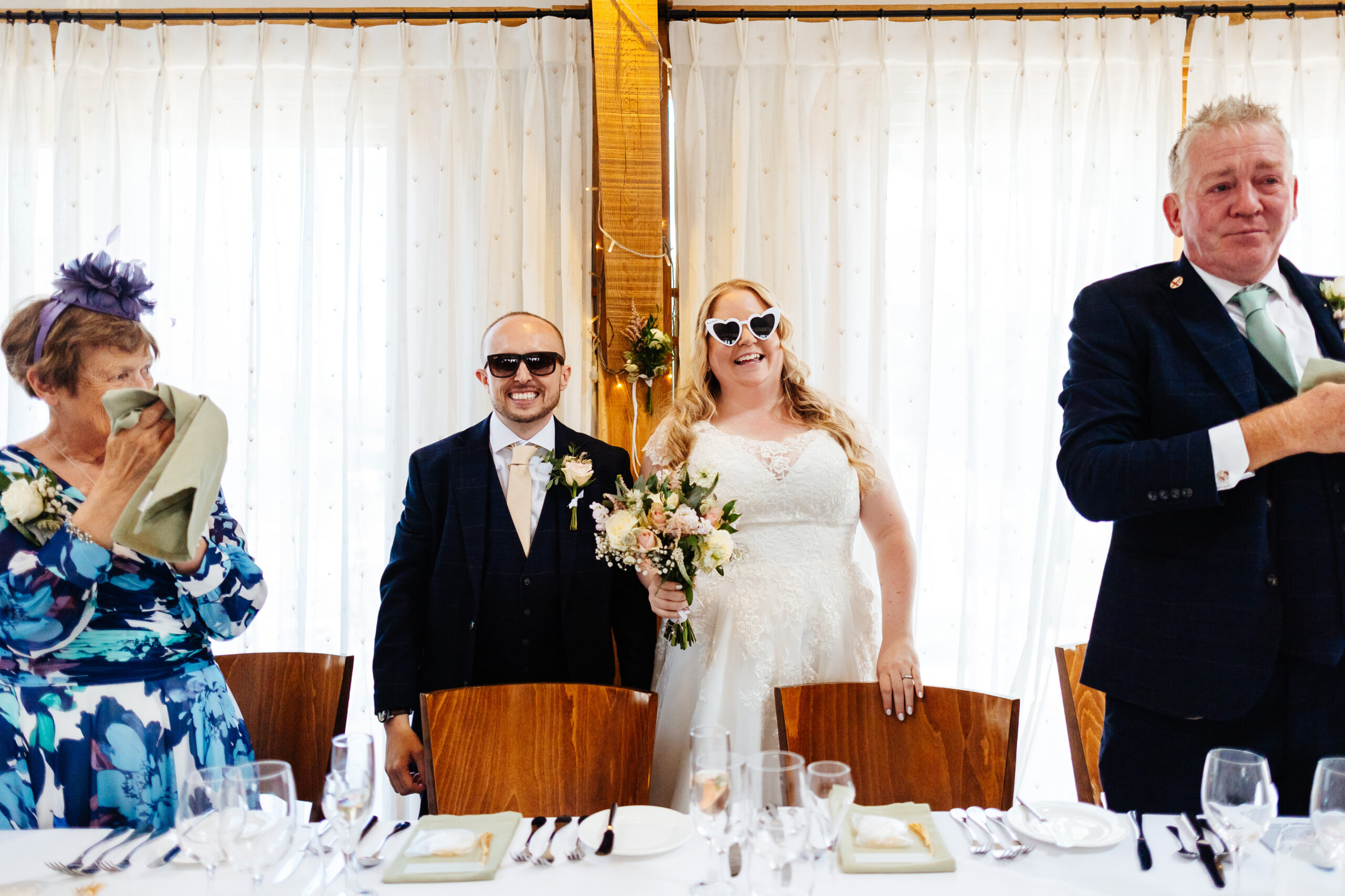 The bride and groom standing at the table. They are wearing sunglasses; the bride's pair are white and heart shaped.