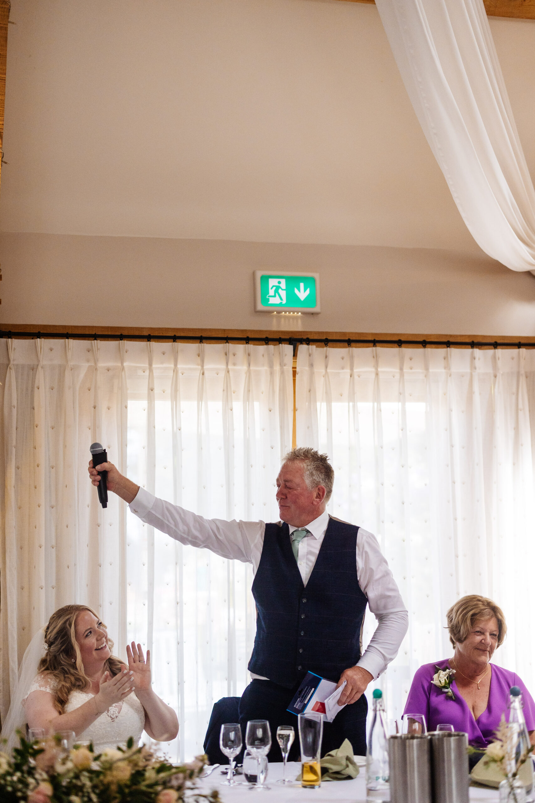 The father of the bride at the table holding up a microphone and looking at his daughter. She is looking at him and clapping.