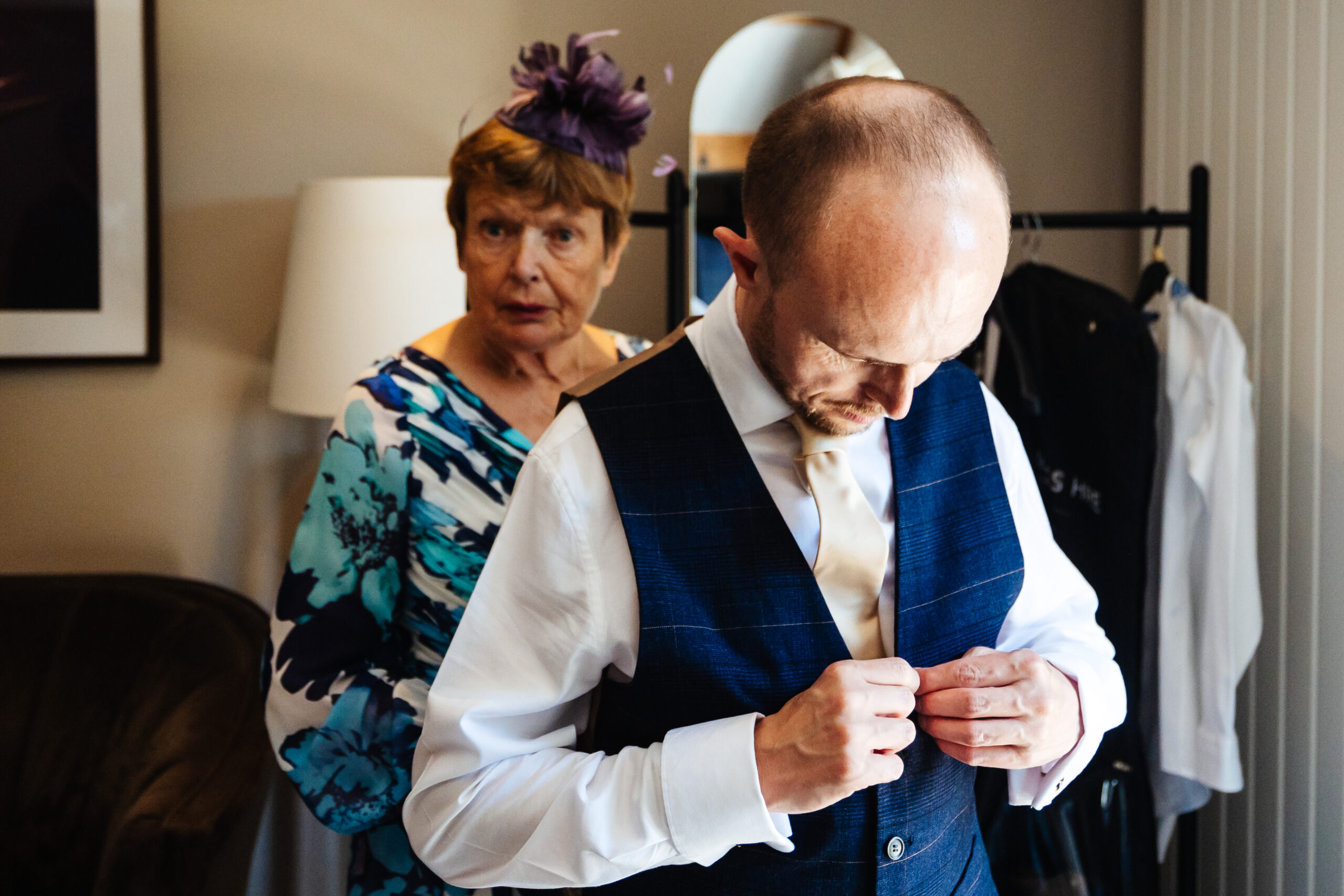 The groom putting his navy blue, striped waistcoat on. He is looking down at the buttons and doing it up with both hands.