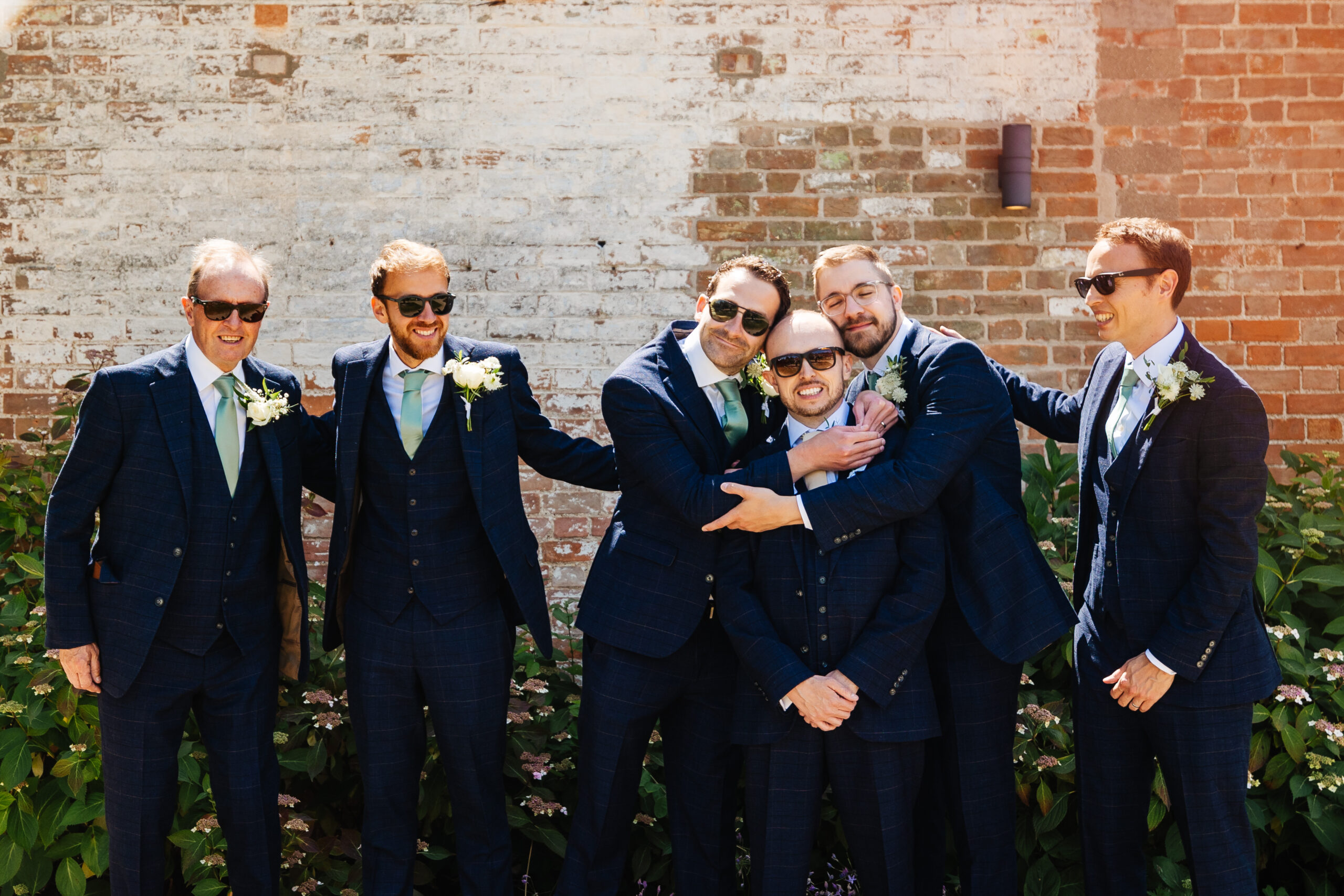 The groom's party stood outside a brick wall. Three of the men are embracing and they are all smiling and having a good time. It is a sunny day and they are all wearing sunglasses.