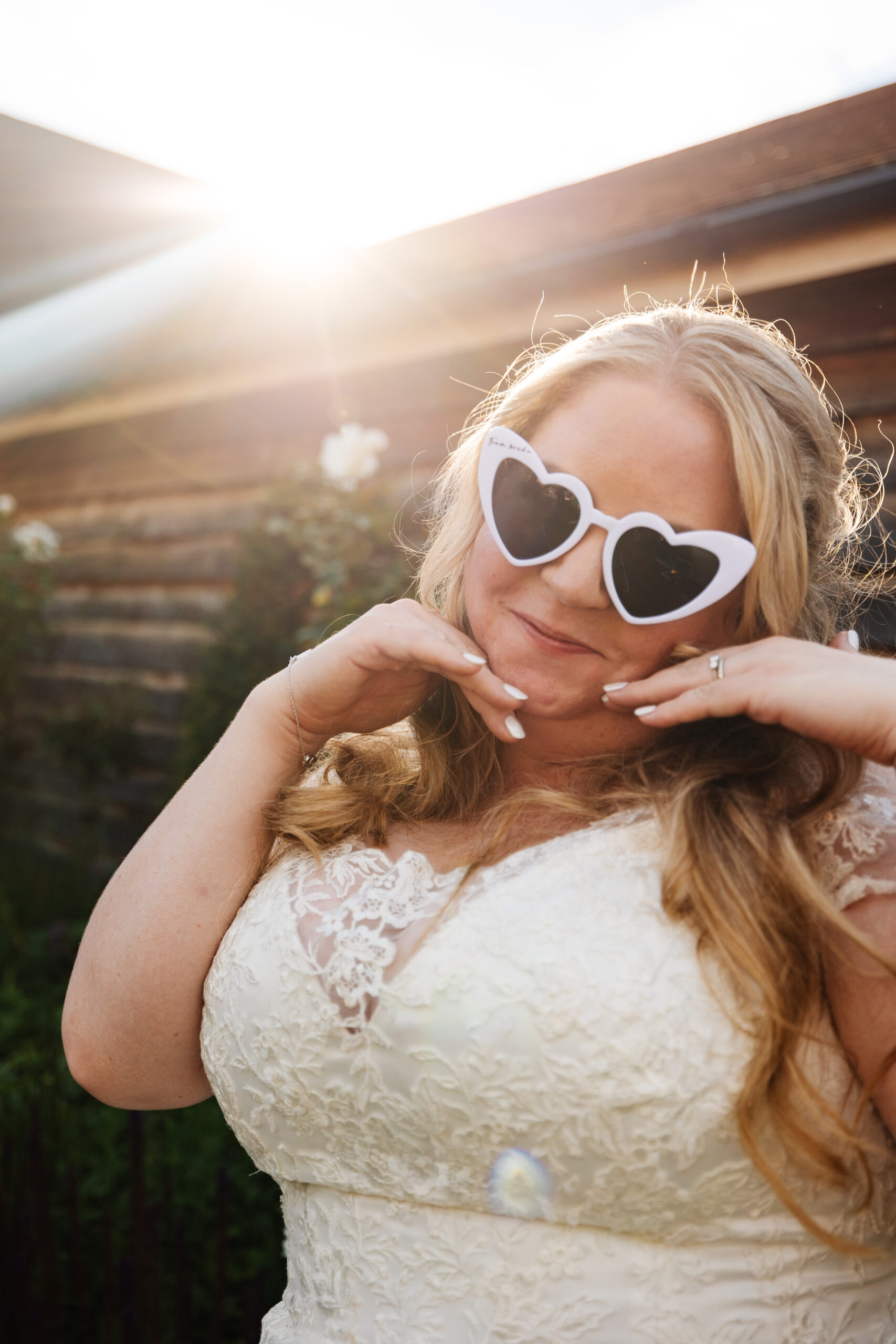 The bride wearing her white, heart sunglassess. She has her hands near her face and is smiling. The sun is beaming in the background.