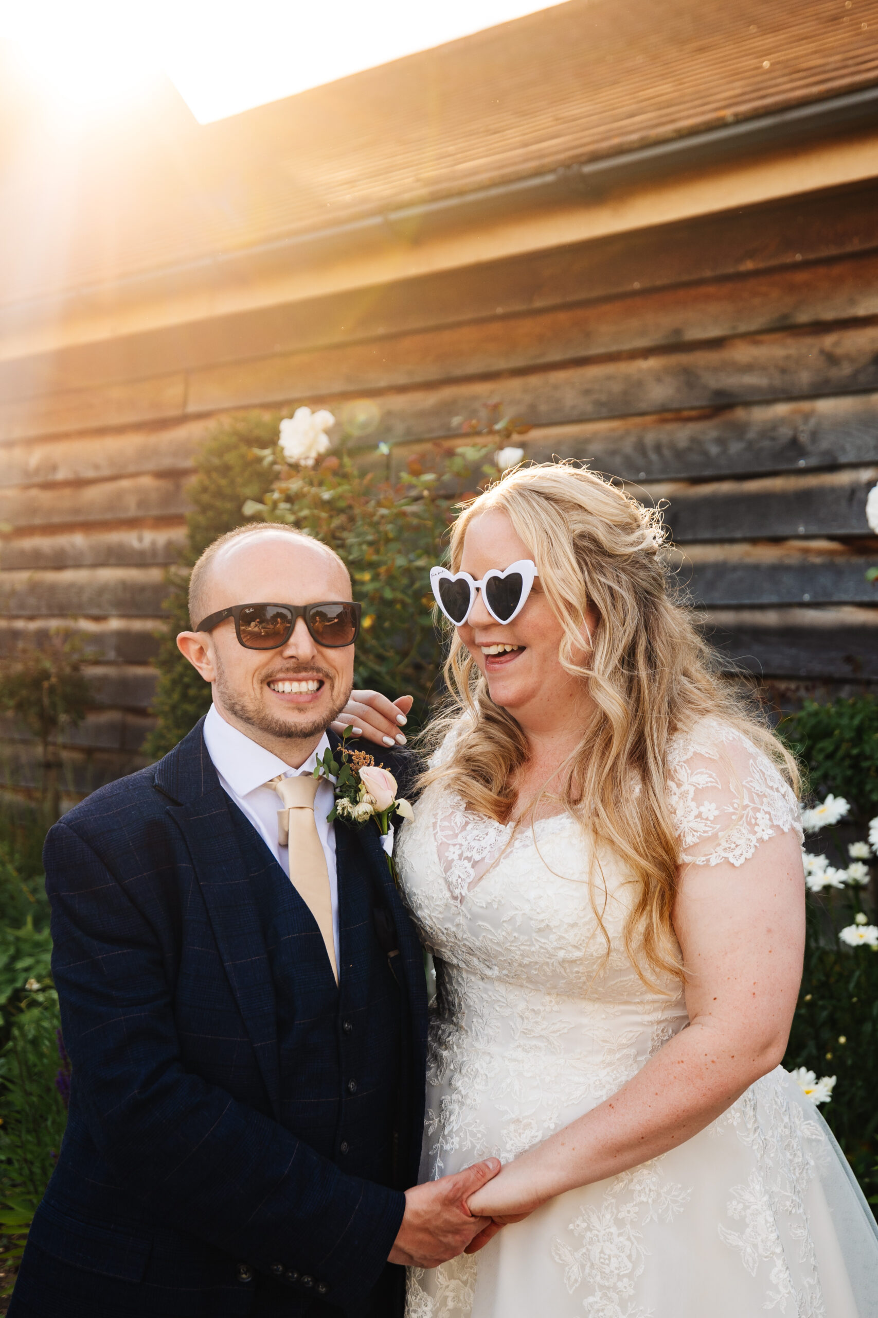 The bride and groom, outside, smiling. The bride has her hand on the groom's shoulder and they are wearing sunglasses. You can see the reflection of the sun in the background.