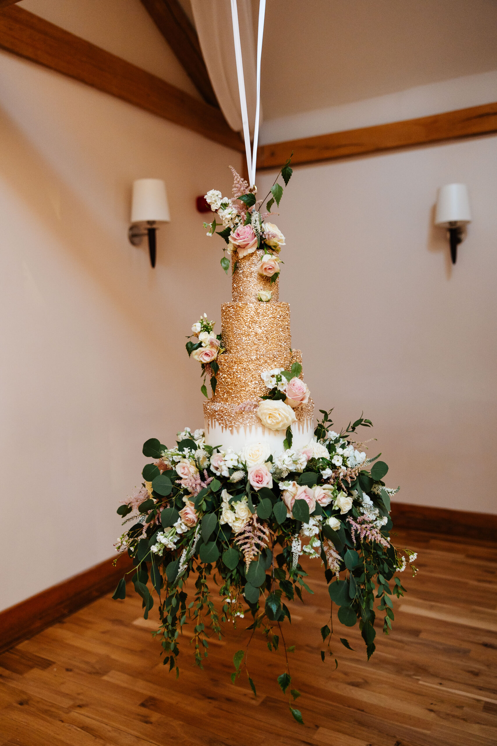 The cake! This is a four tiered cake with the first three layers covered in gold glitter. The bottom tier is white with some gold glitter on and it has flowers surrounding the bottom. It is suspended from the ceiling!!!