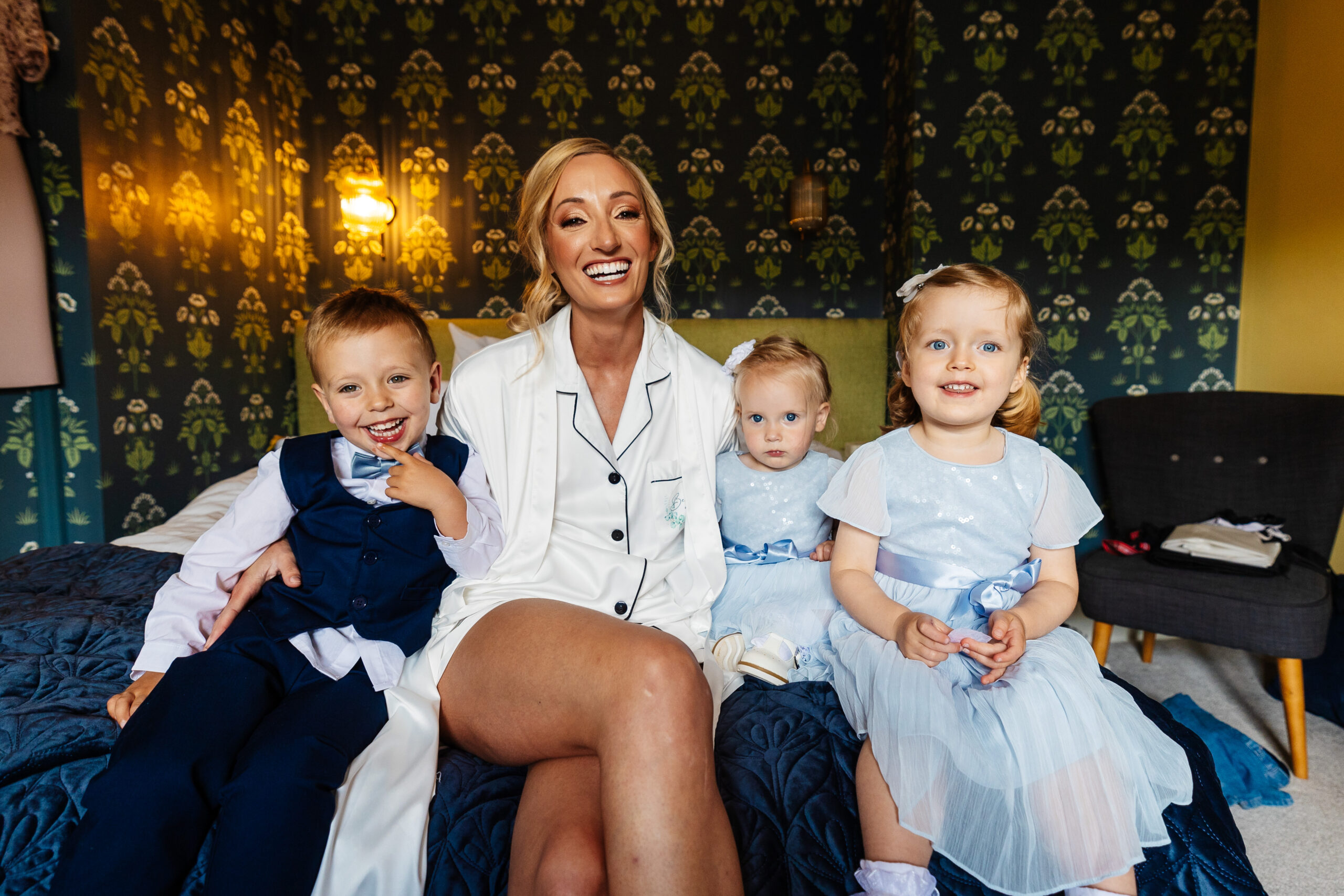 A bride dressed in white silk pajamas with a black lace. She is sat on a bed and has three children next to her. She has her arm around a little boy and a little girl. The boy has a dark navy suit on with a white shirt and the girls are wearing pale blue dresses with ribbons around them.