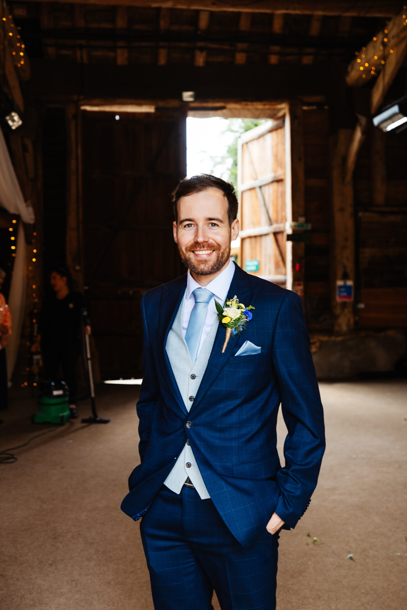 The groom dressed in a dark navy suit, standing in a barn and smiling at the camera.