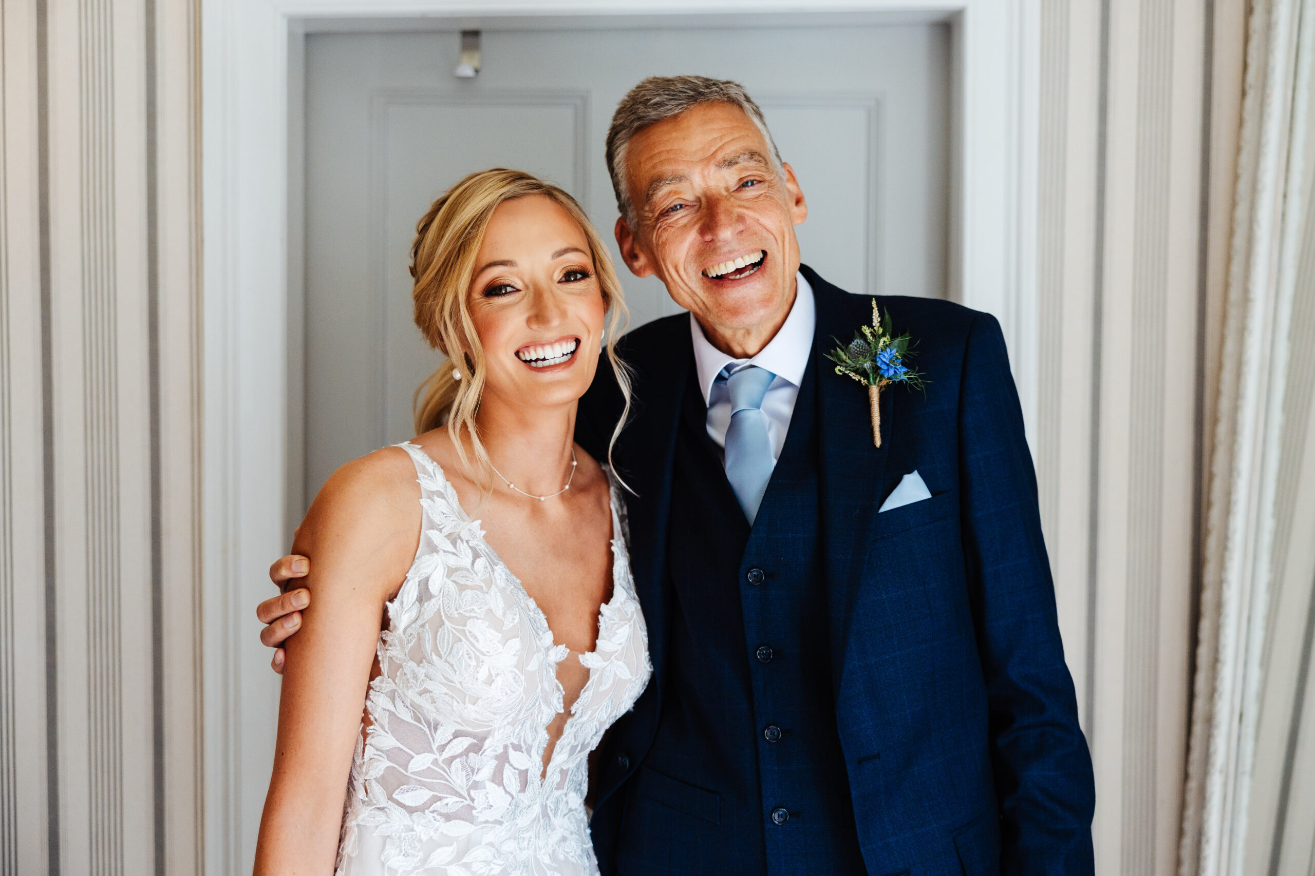 A bride in a lacey white dress and her father in a dark navy suit. He has his arm around her and they are looking at the camera and smiling.