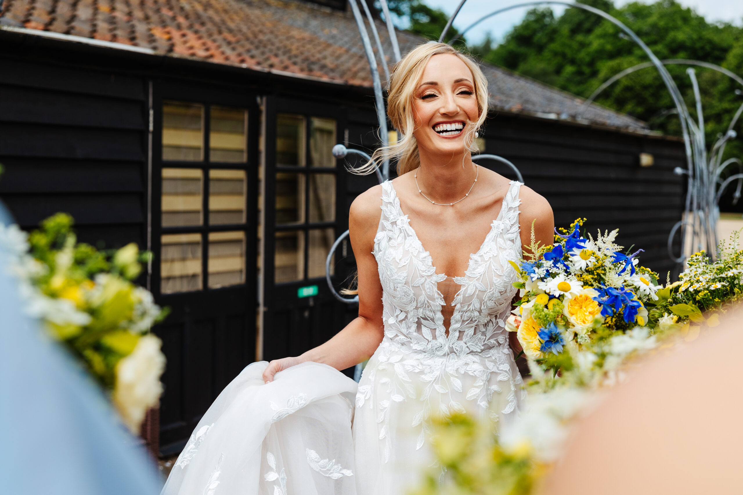 A bride wearing a skinny strapped lacey dress. She is holding the bottom of her dress in one hand and a bunch of flowers in the other and has a huge smile on her face.