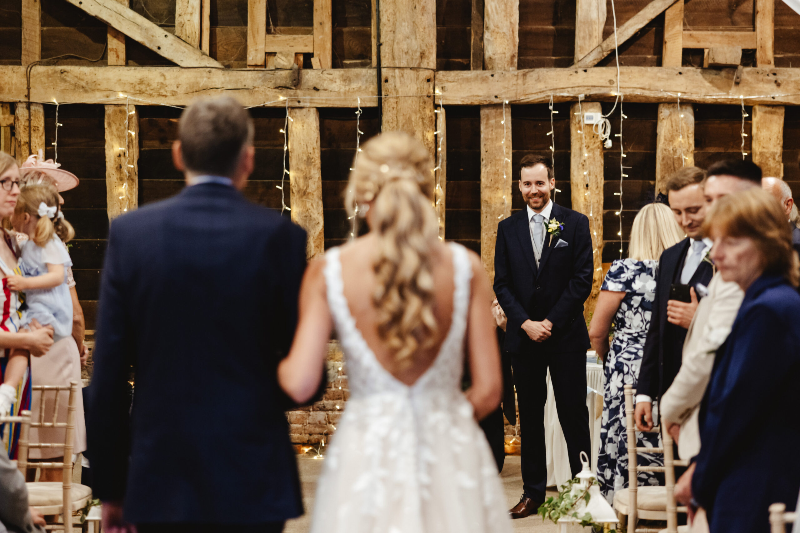 An image inside a barn, you can see the bride and her father's backs and you can see the groom stood at the alter smiling.