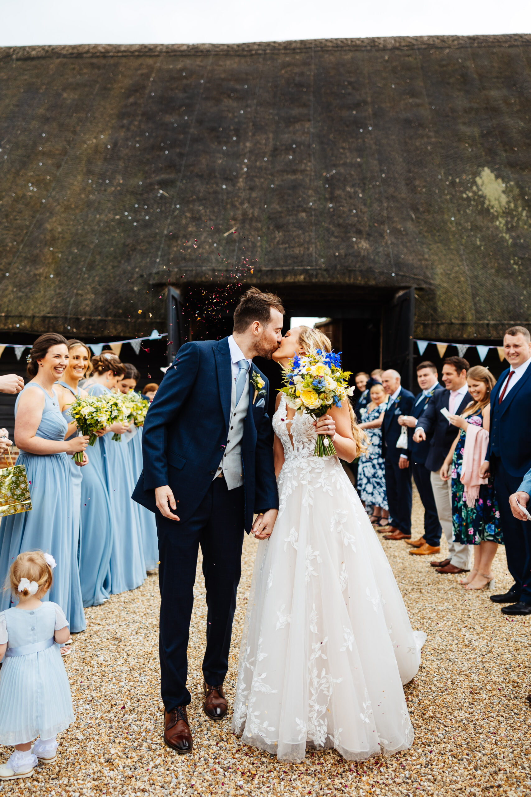 A bride and groom outside surrounded by their friends and families who are smiling and looking at them. The bride and groom are kissing and the bride is holding a big bunch of yellow and blue flowers.