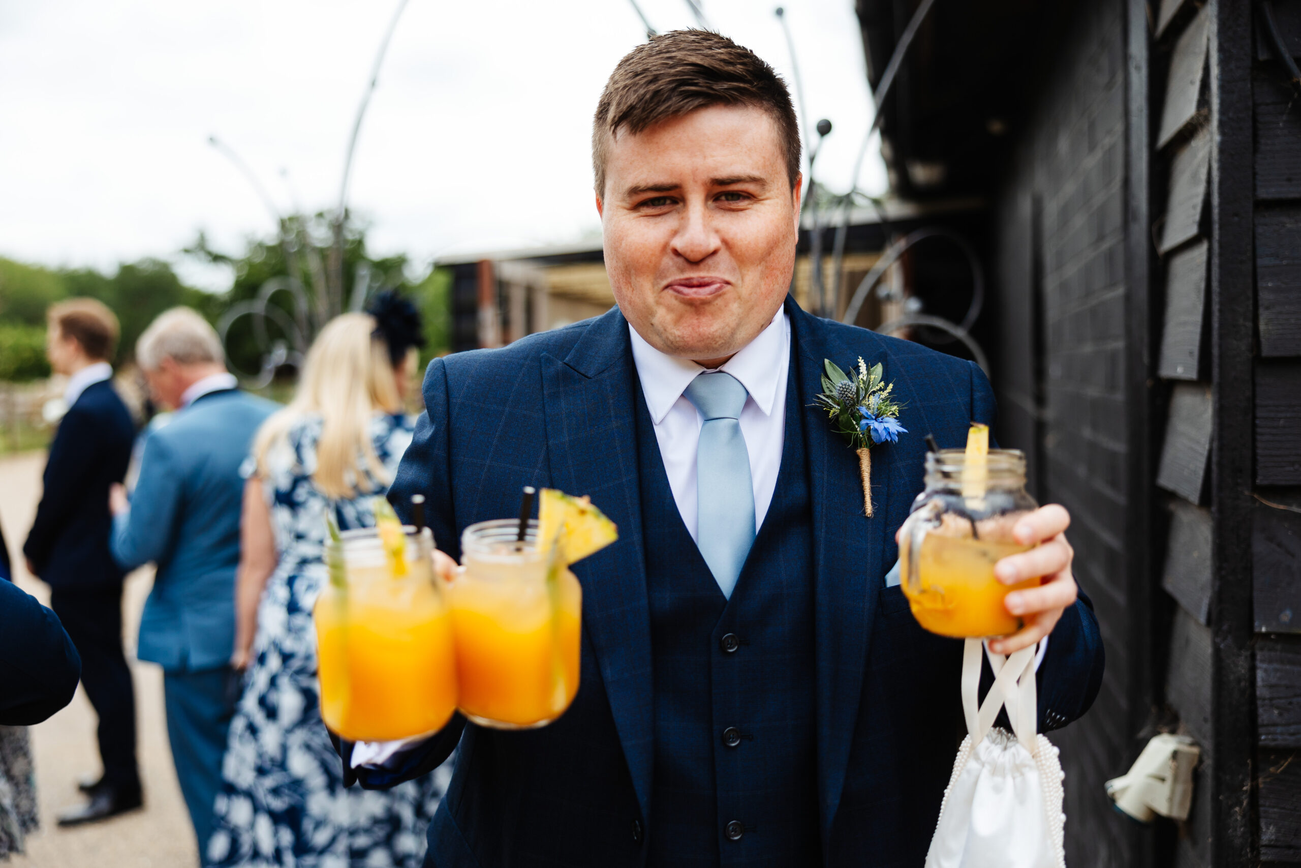 A man in a dark navy suit holding three bright orange drinks. He has a cheeky look on his face.