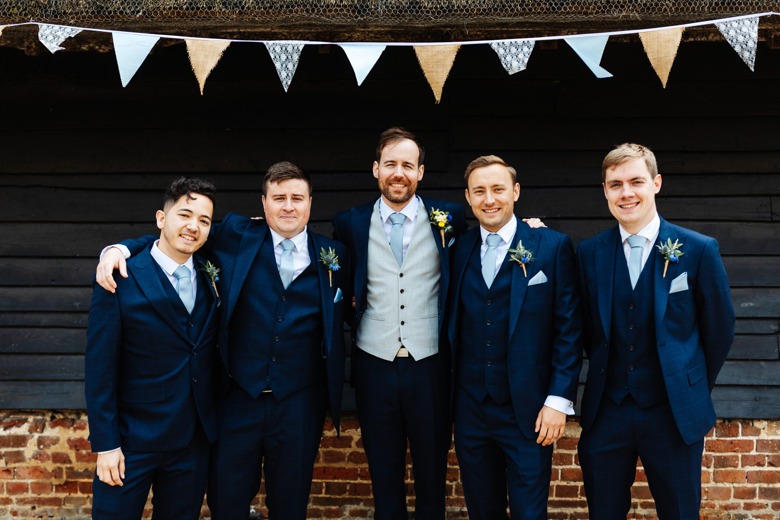 A group of men dressed in dark navy blue suits looking at the camera smiling. They are stood in front of a barn and colourful bunting above them