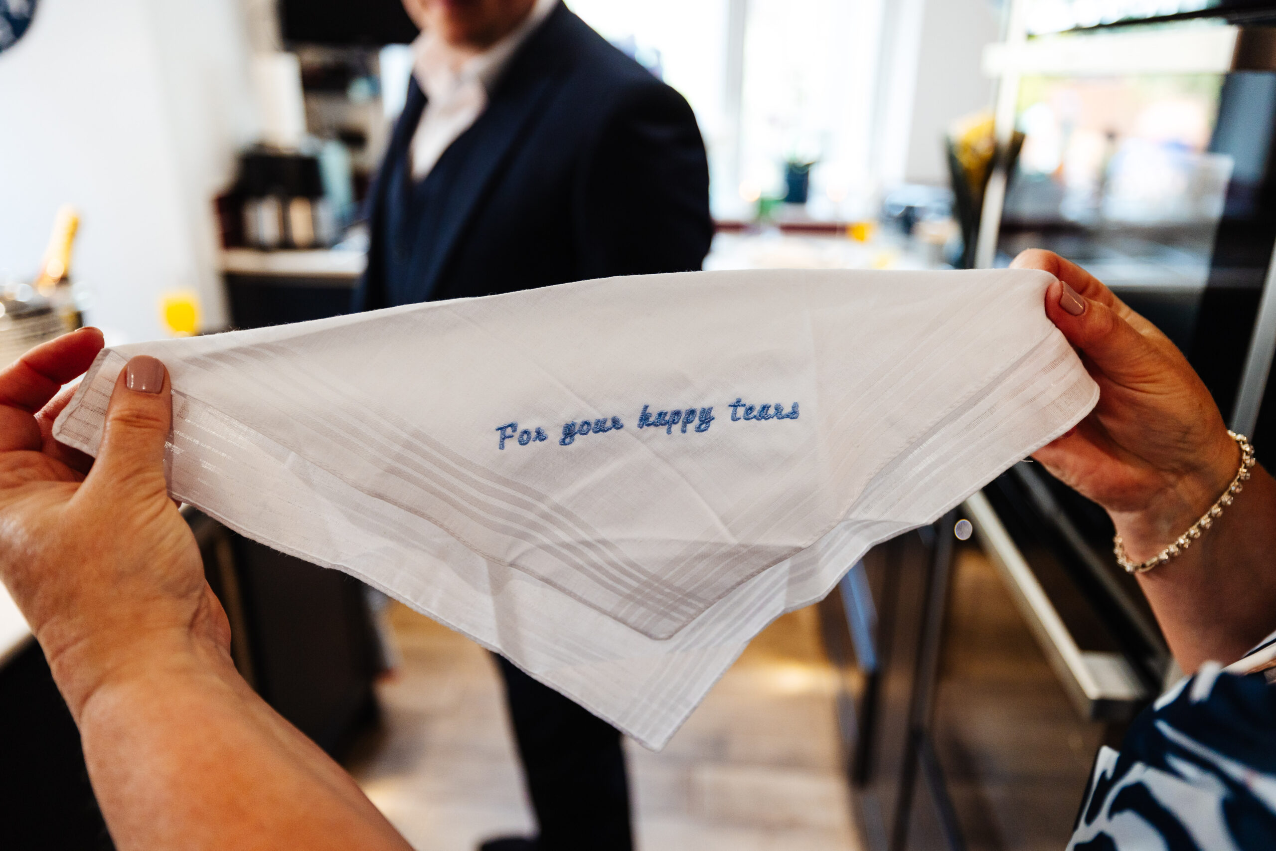 A pair of hands holding a napkin which is embroidered with "for your happy tears" in blue stitching.