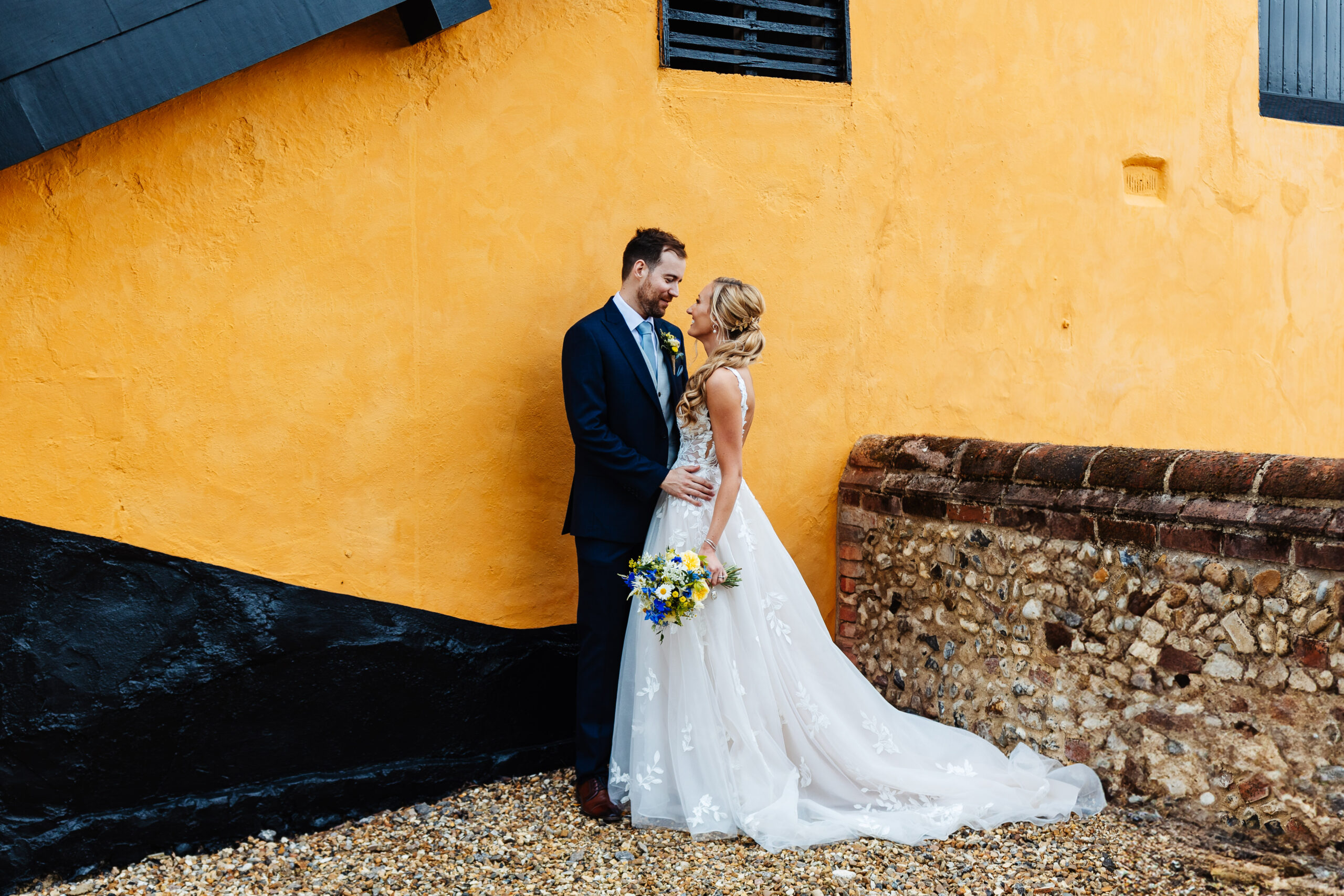 A bride and groom stood in front of a yellow building. They are looking in each other's eyes