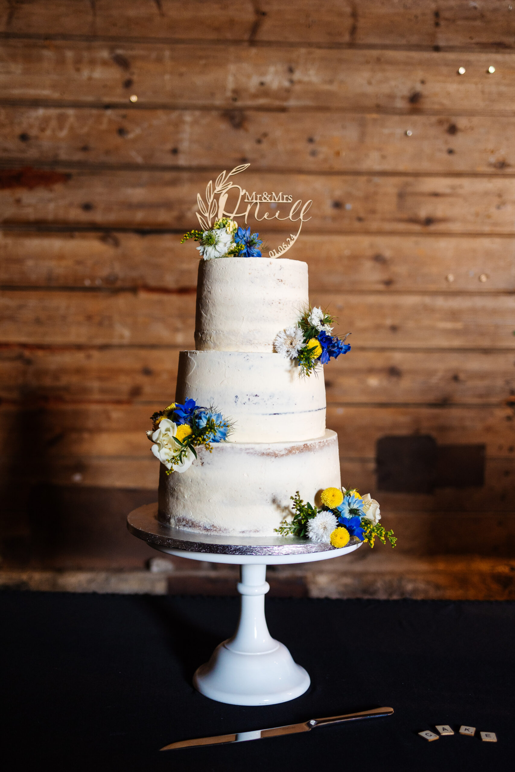 A three tiered wedding cake which is covered in white frosting and has blue and yellow flowers on it. The top sign says Mr & Mrs O'Neill in a wooden circular sign and the date of their wedding on too.