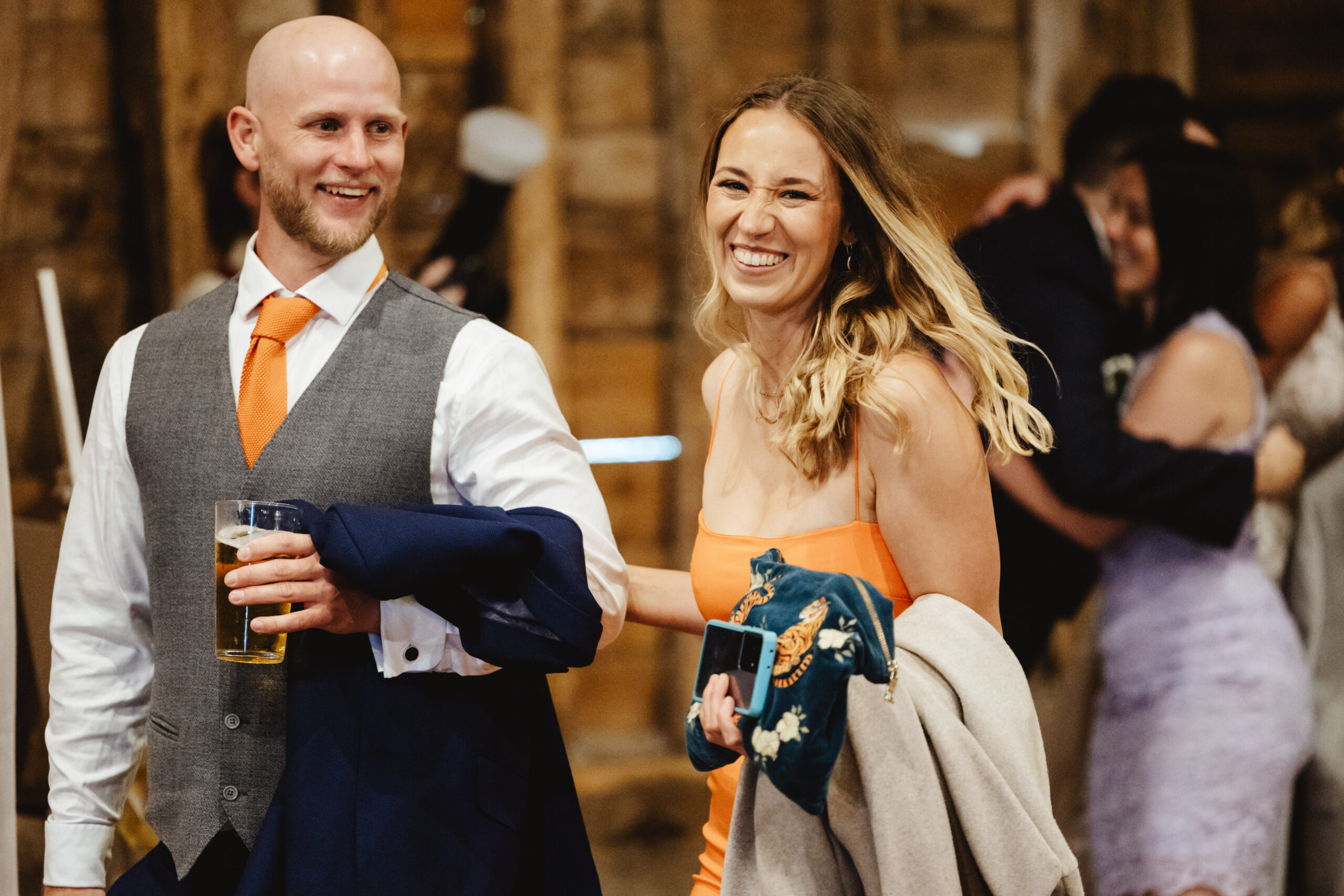 A man and a woman guest smiling. The lady is wearing an orange dress holding a blue clutch bag and the man is wearing a white shirt, grey waistcoat and orange tie. He is holding a beer in one hand. She is holding his arm.