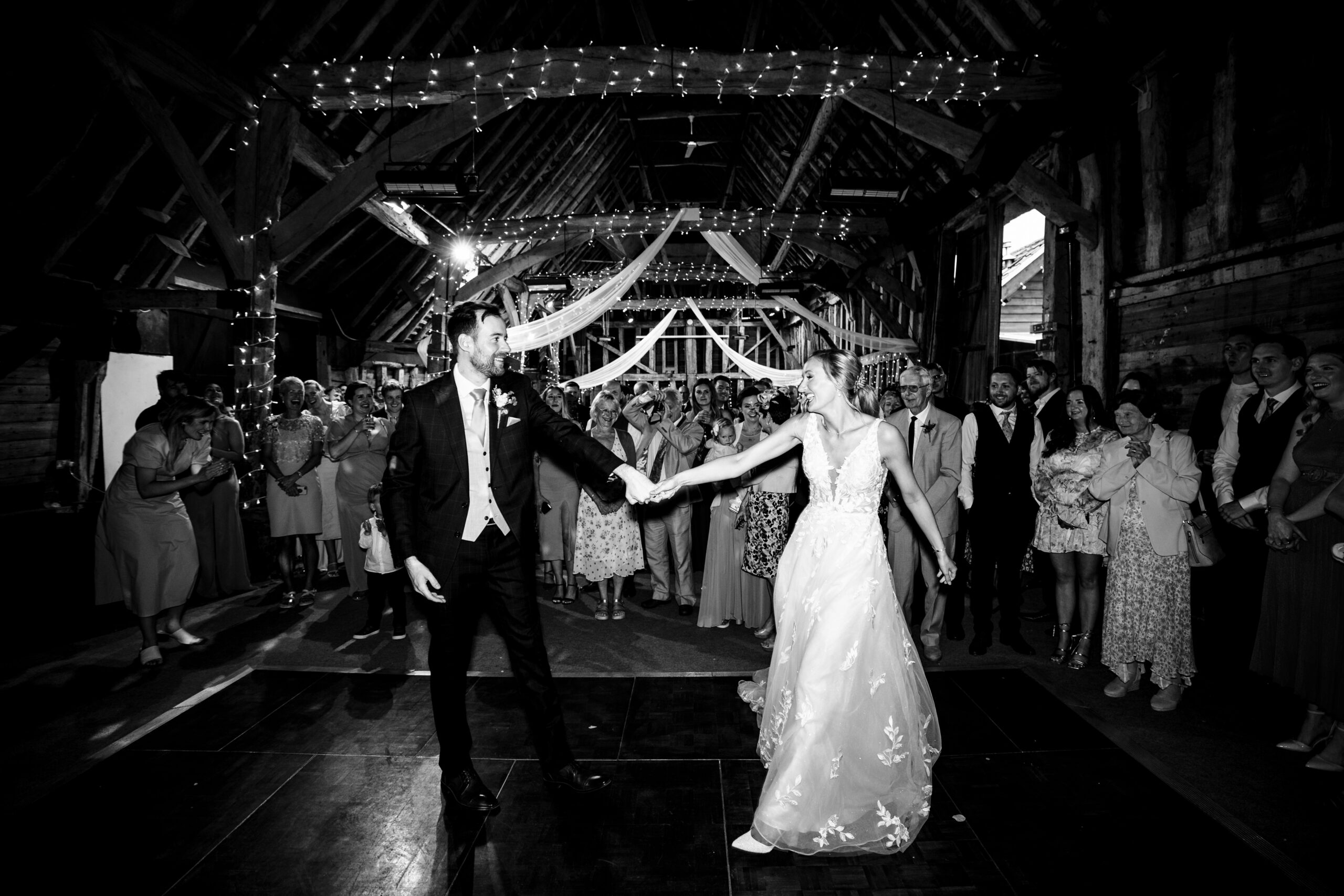 A black and white image of a bride and a groom on a dance floor surrounded by their friends and family. They are holding hands.