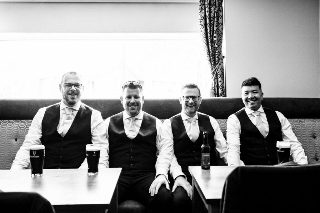 A black and white image of four males sitting on a pub seat with drinks in front of them. They are all looking at the camera and smiling.