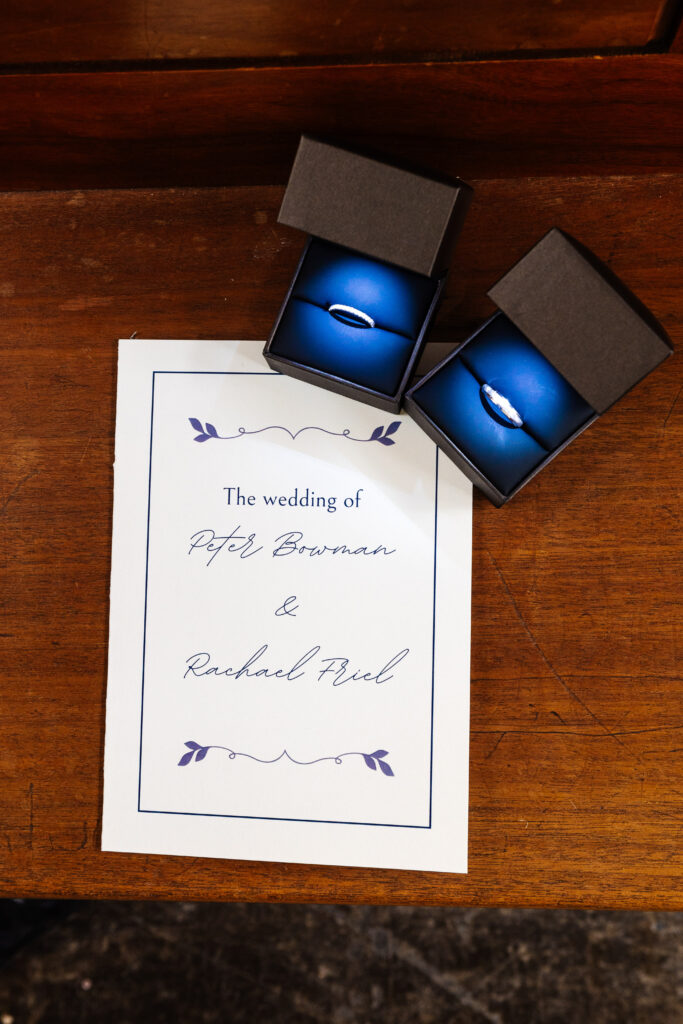 An image of two rings in their boxes. The lids are open and show a bright blue inside. The rings are silver. There is also the order of ceremony on a wooden table.