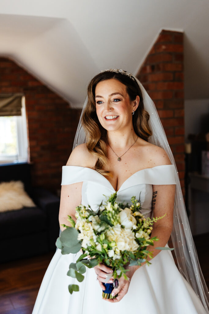 A bride looking to the side in her white off the shoulder dress. She has a veil in and is holding her flowers. She is smiling.