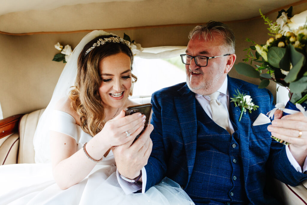 A bride and her father in the back of a car. He is showing her his phone and holding her flowers for her. She is laughing.