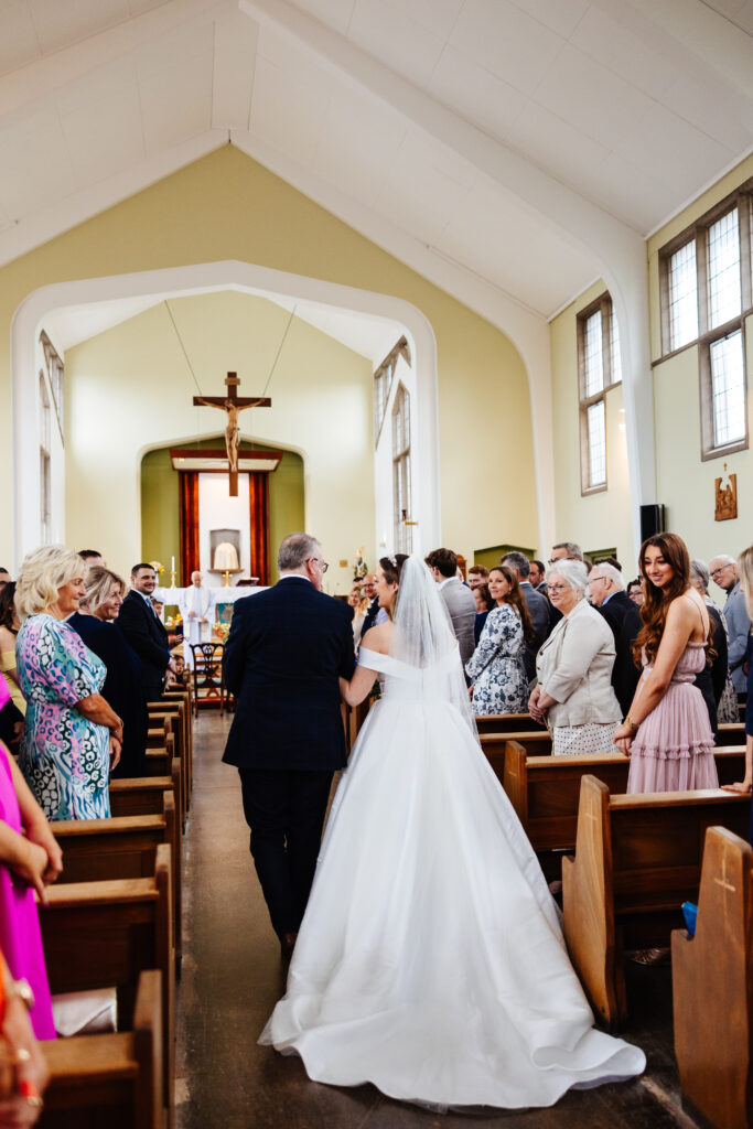 A man in a black suit and a woman in a long flowing white wedding dress and veil. They are walking down an aisle of a church with their friends and family by their sides.