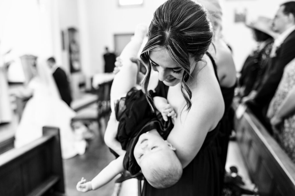 A black and white image of a woman holding a baby upside down in a playful way in a church. You can see the bride and groom in the photo