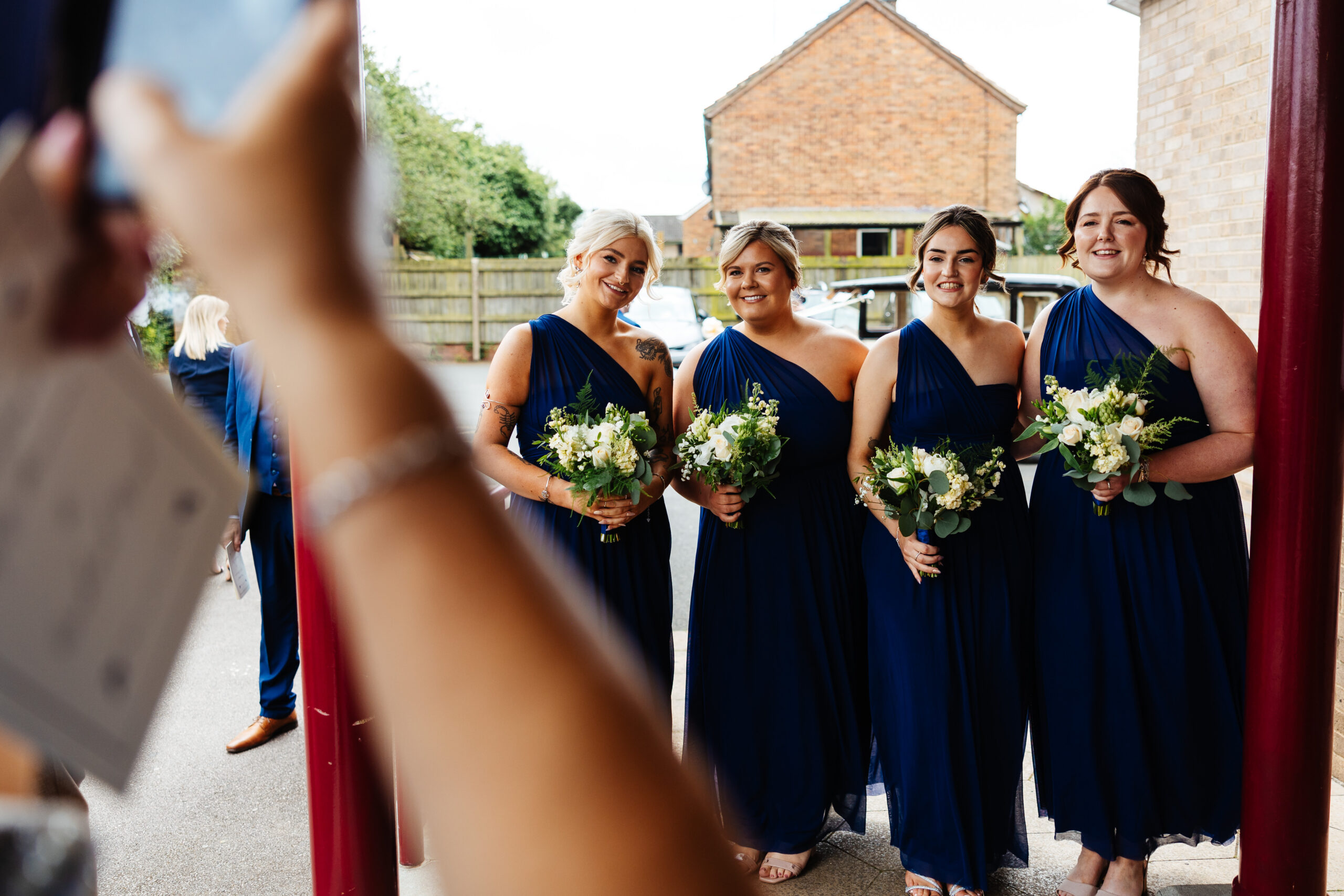 Four bridesmaids wearing navy blue one shouldered dresses. They are holding a bunch of white flowers, roses to be specific. Someone is taking their photograph and they are also in this picture.