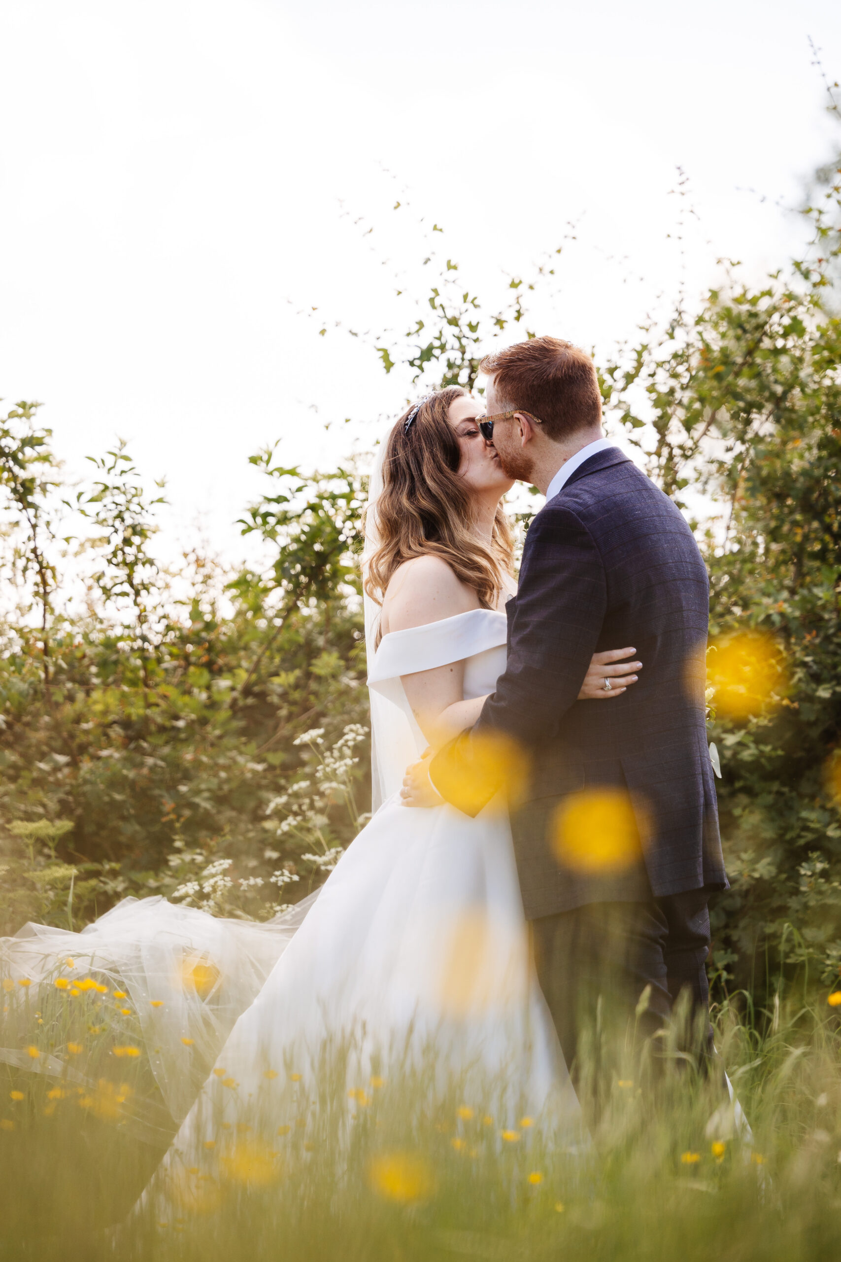 A bride and groom looking at each other standing in a luscious green field. They are kissing each other with their eyes closed. The bride is in an off the shoulder white dress and the groom is wearing a navy suit with a white shirt and sunglasses.