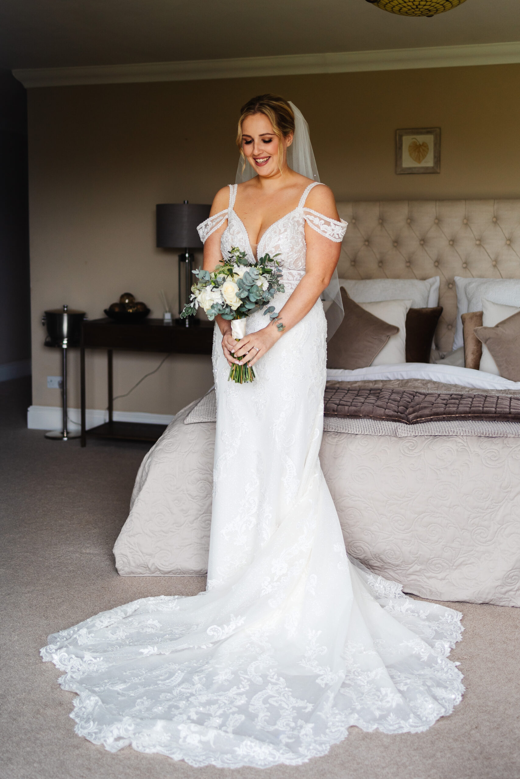 A bride in a long, flowing white lace dress and veil holding a bouquet of flowers. She is looking down and smiling.