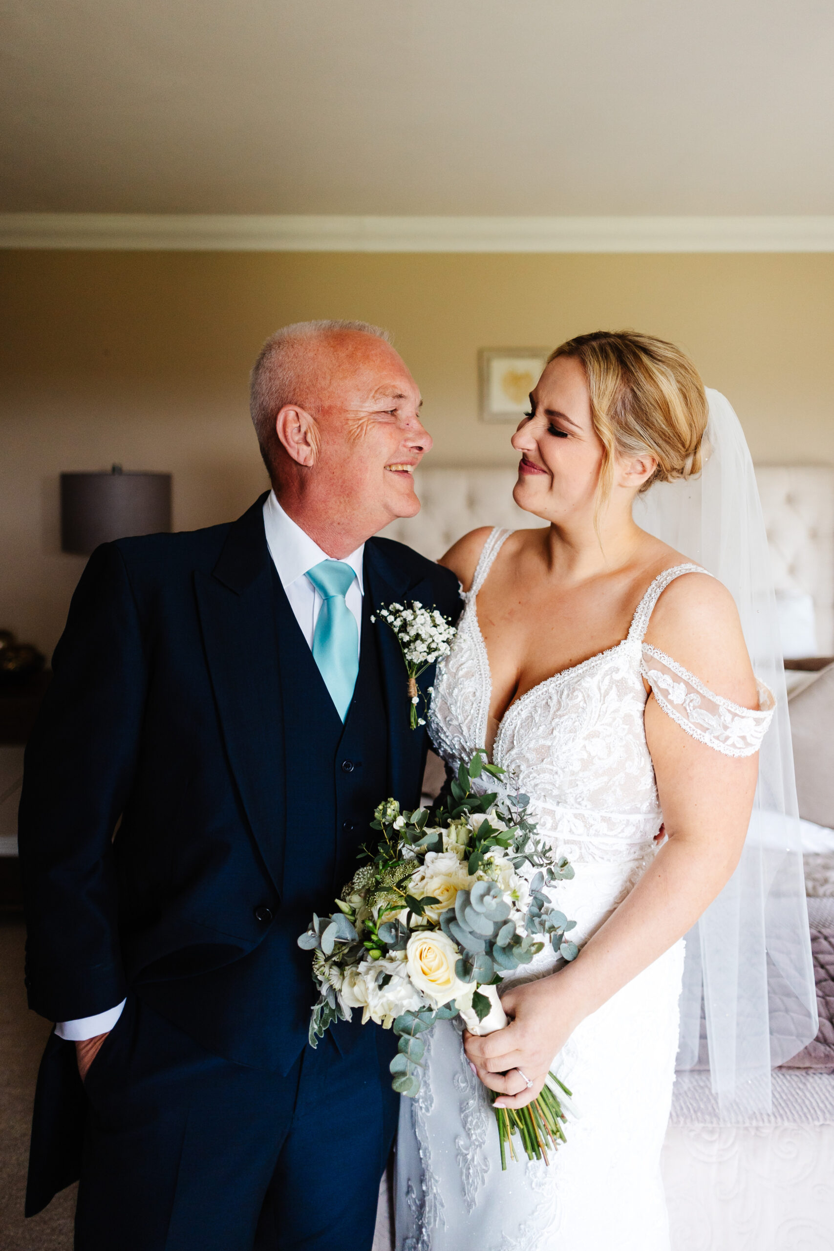 A father and his daughter looking at each other. The lady is in a white wedding dress and veil and the man is in a navy blue suit. They are smiling and the bride has her nose scrunched up.