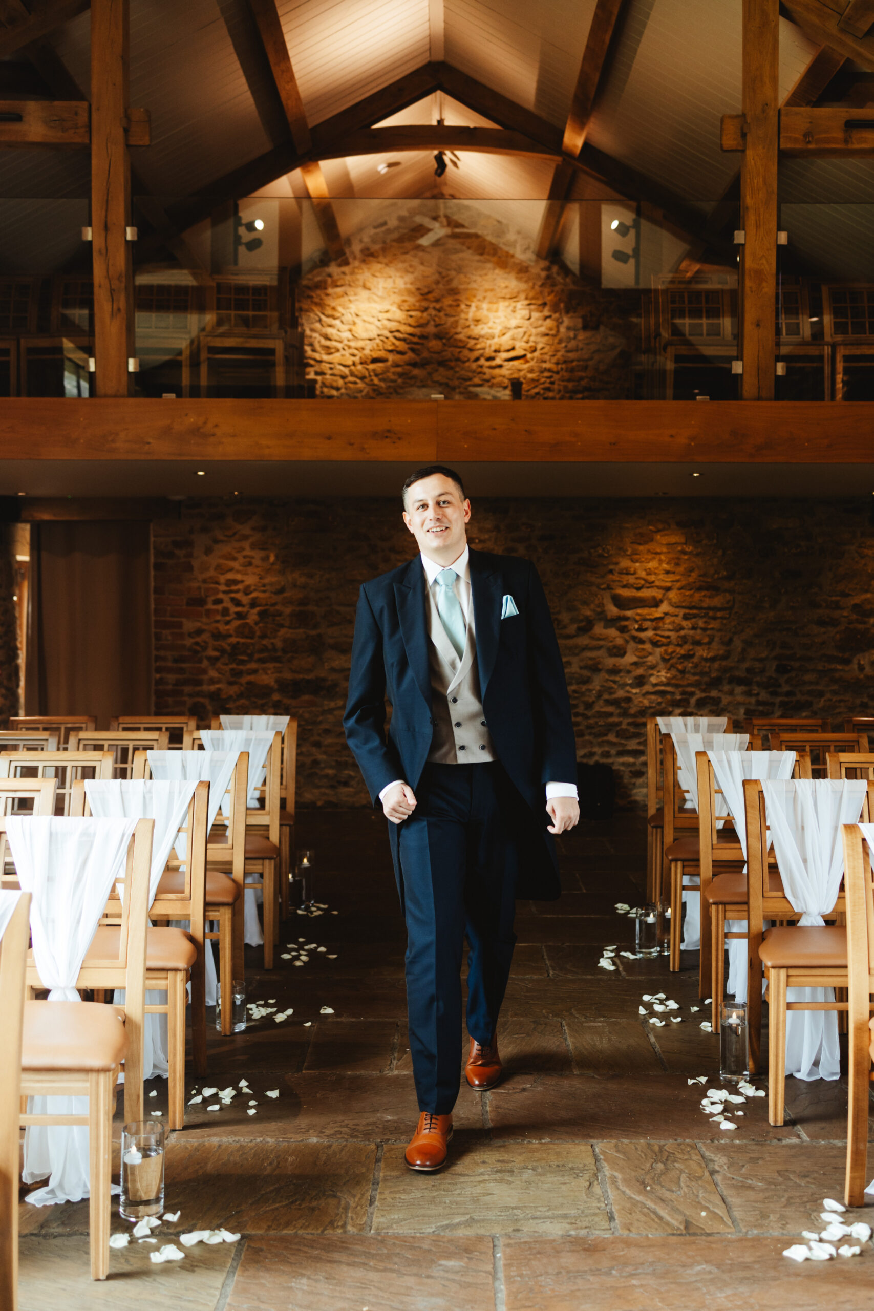 A groom in a navy blue suit and brown shoes walking down the aisle of his wedding venue. He is smiling nerviously.