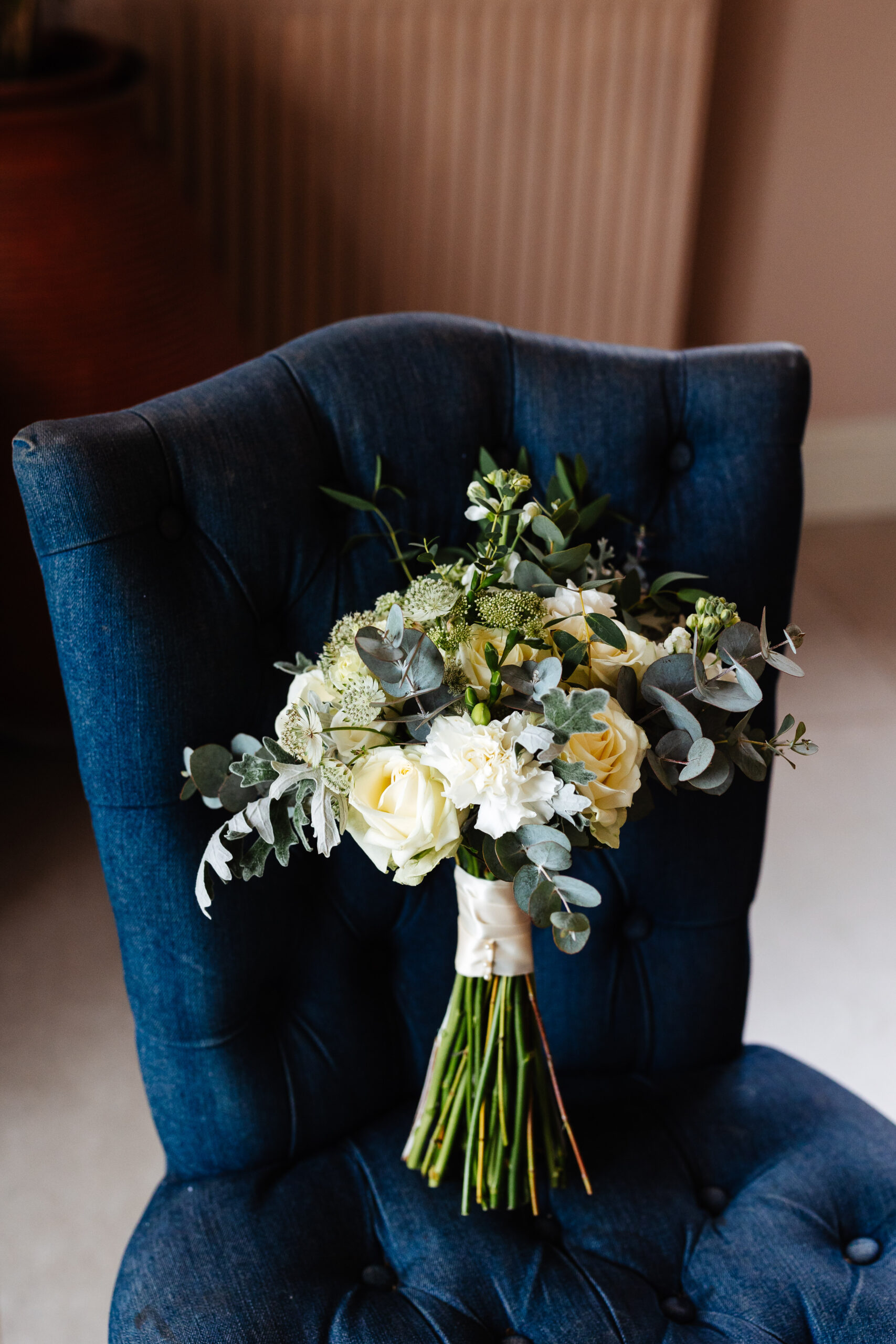 A beautiful bouquet of white roses and eucalyptus propped on a plush, navy blue chair