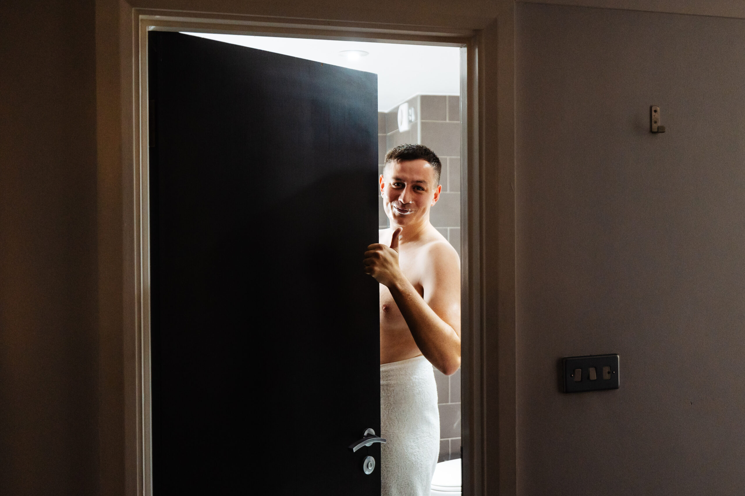 The groom standing in a bathroom doorway. He is wearing a towel and has shaving cream on his face.