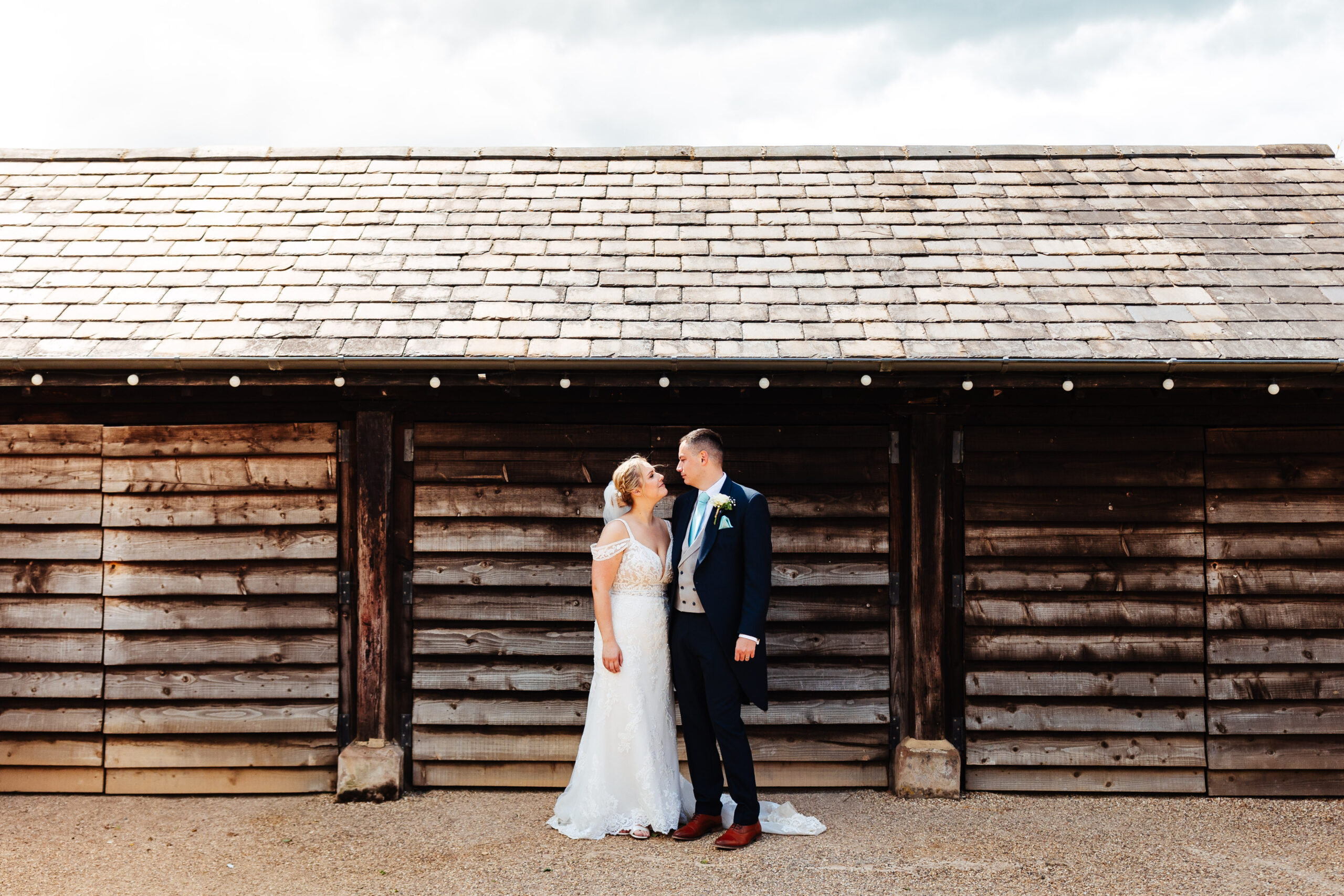 A bride and groom standing in front of a barn. They are smiling at each other.