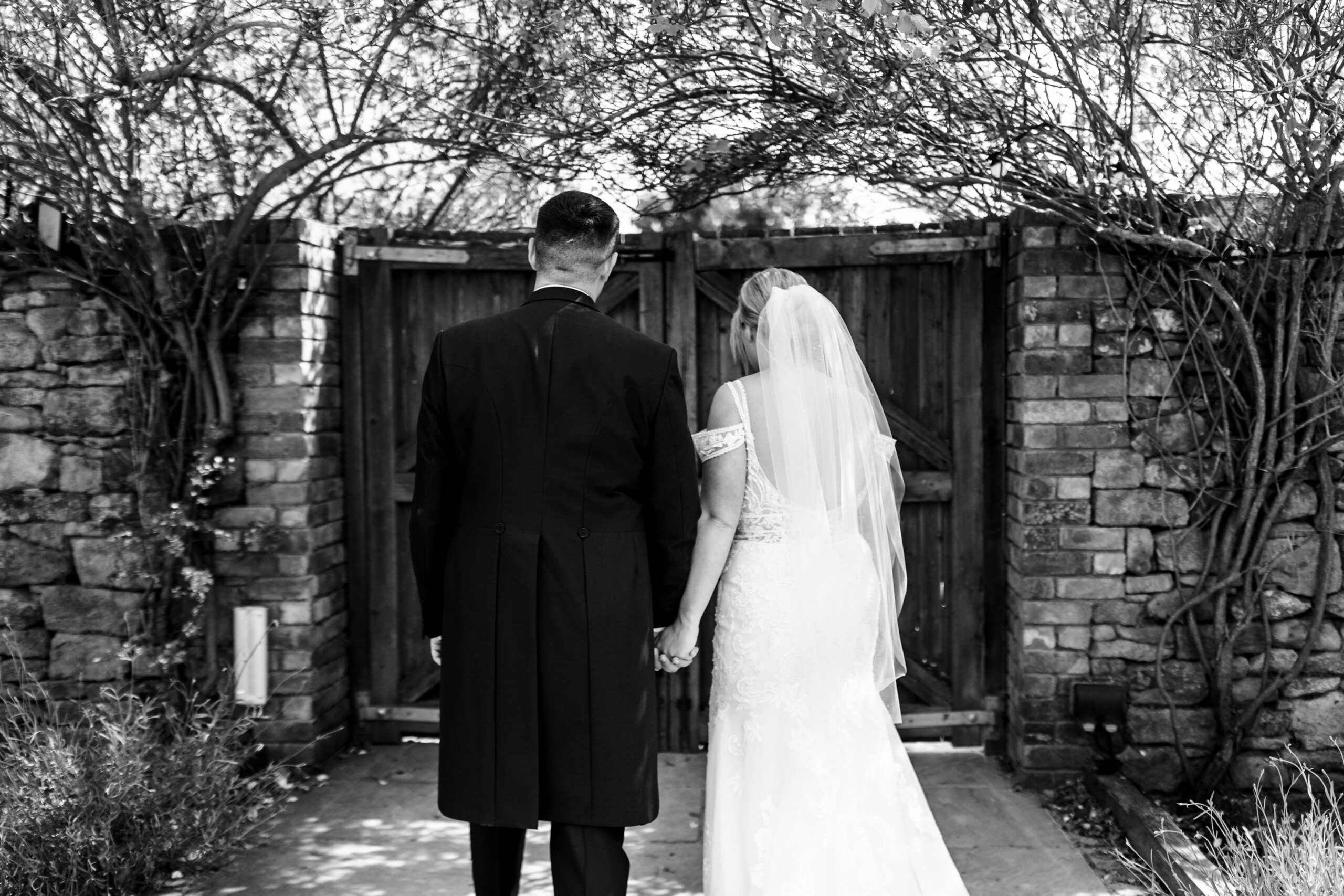 The back of a bride and groom. The photo is black and white. They are standing outside a gate between two brick walls.