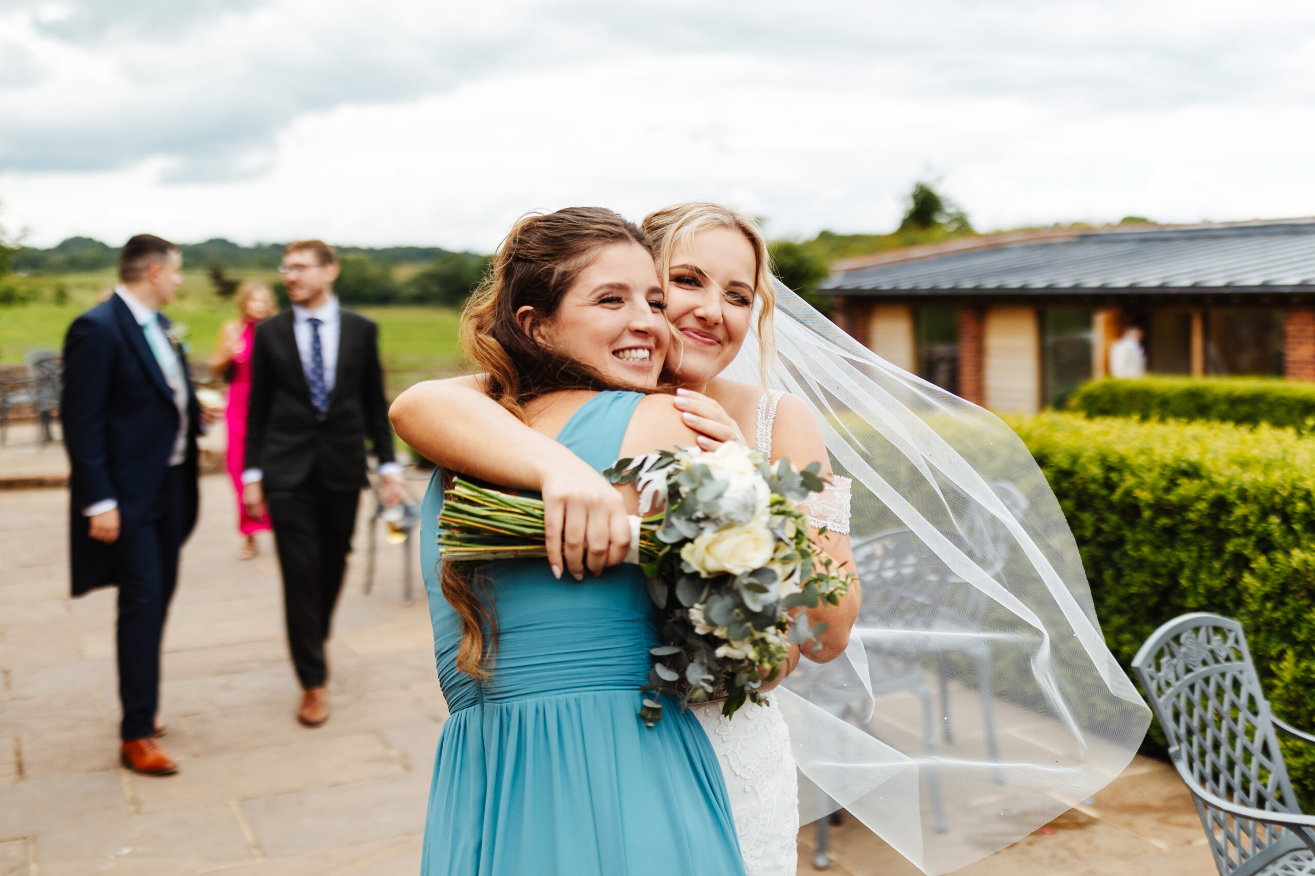 A bride hugging her bridesmaid. They are both smiling and the bride's veil is floating in the wind.