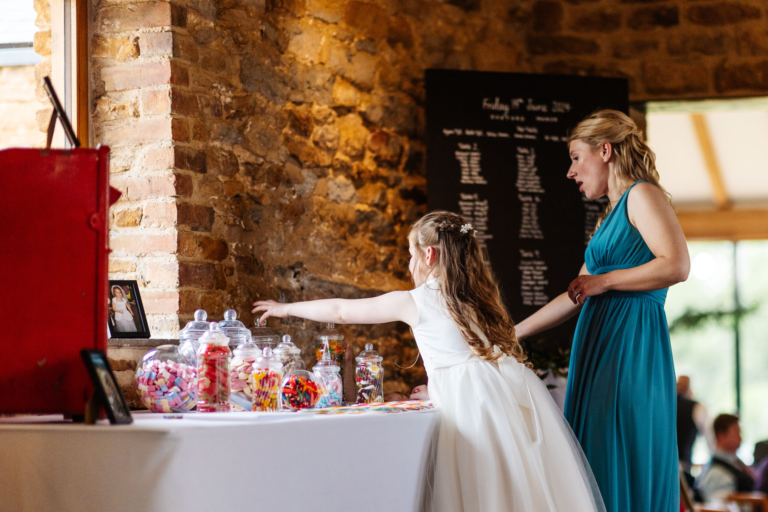 A little girl in a long white dress reaching for the sweetie table! There is a bridesmaid helping her. The sweets are in glass jars and look colourful.