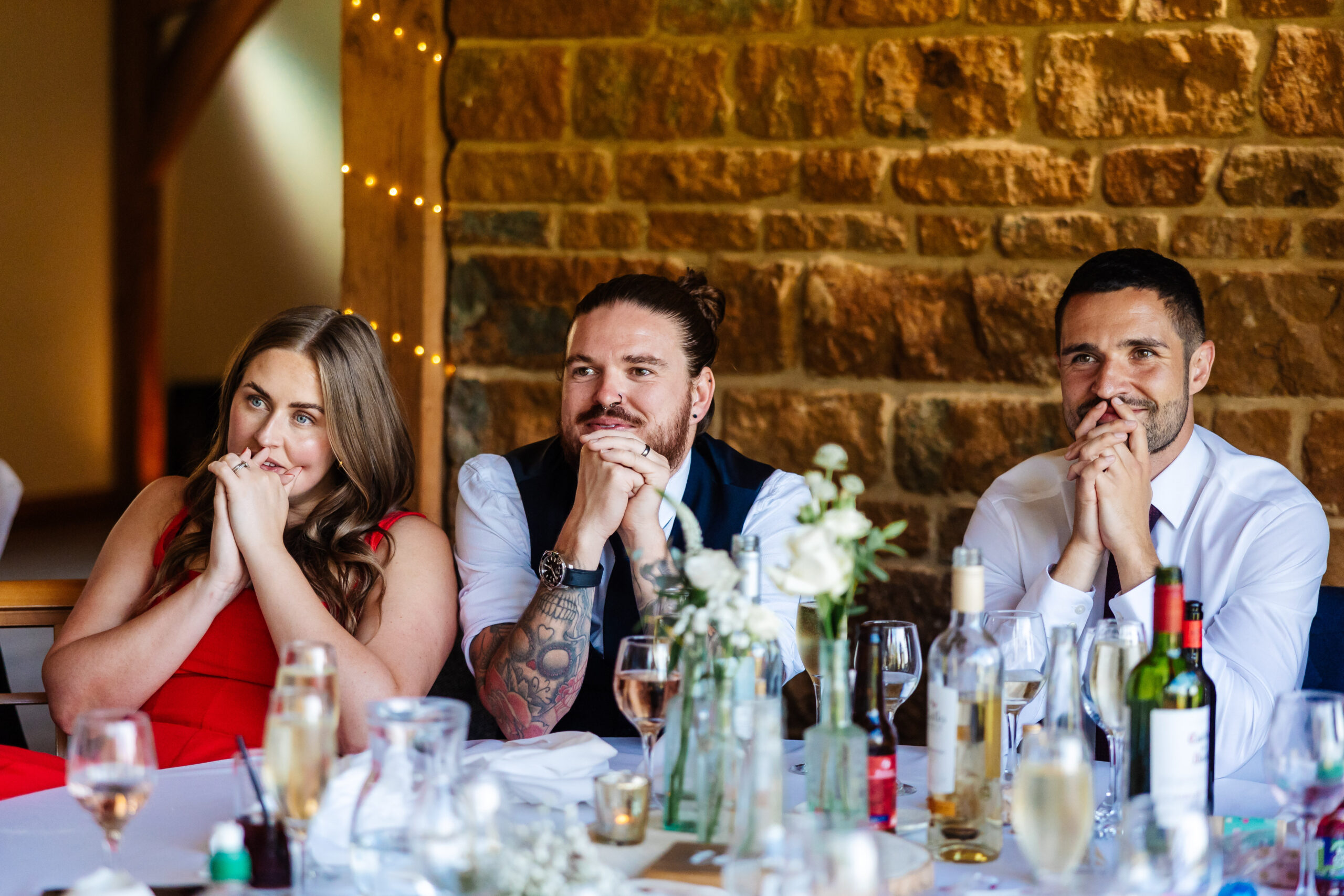 Three wedding guests looking at the bride and groom. They are sat at a table and smiling as they listen to the wedding speeches.