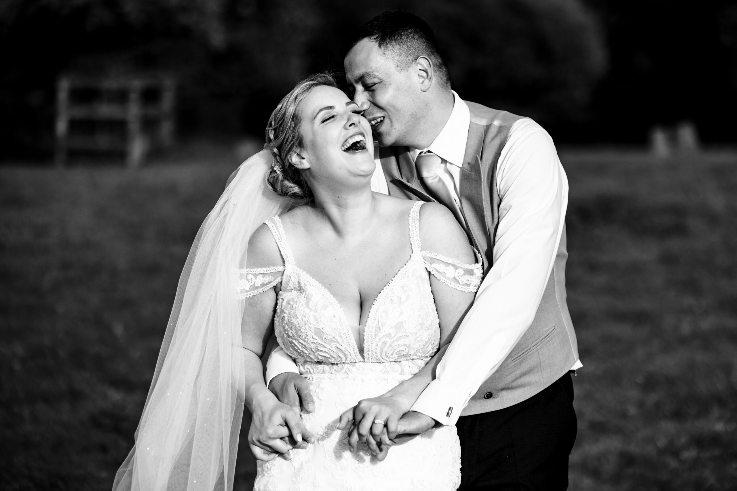 A black and white image of a bride and groom. The groom is hugging the bride from behind and they are smiling and laughing.