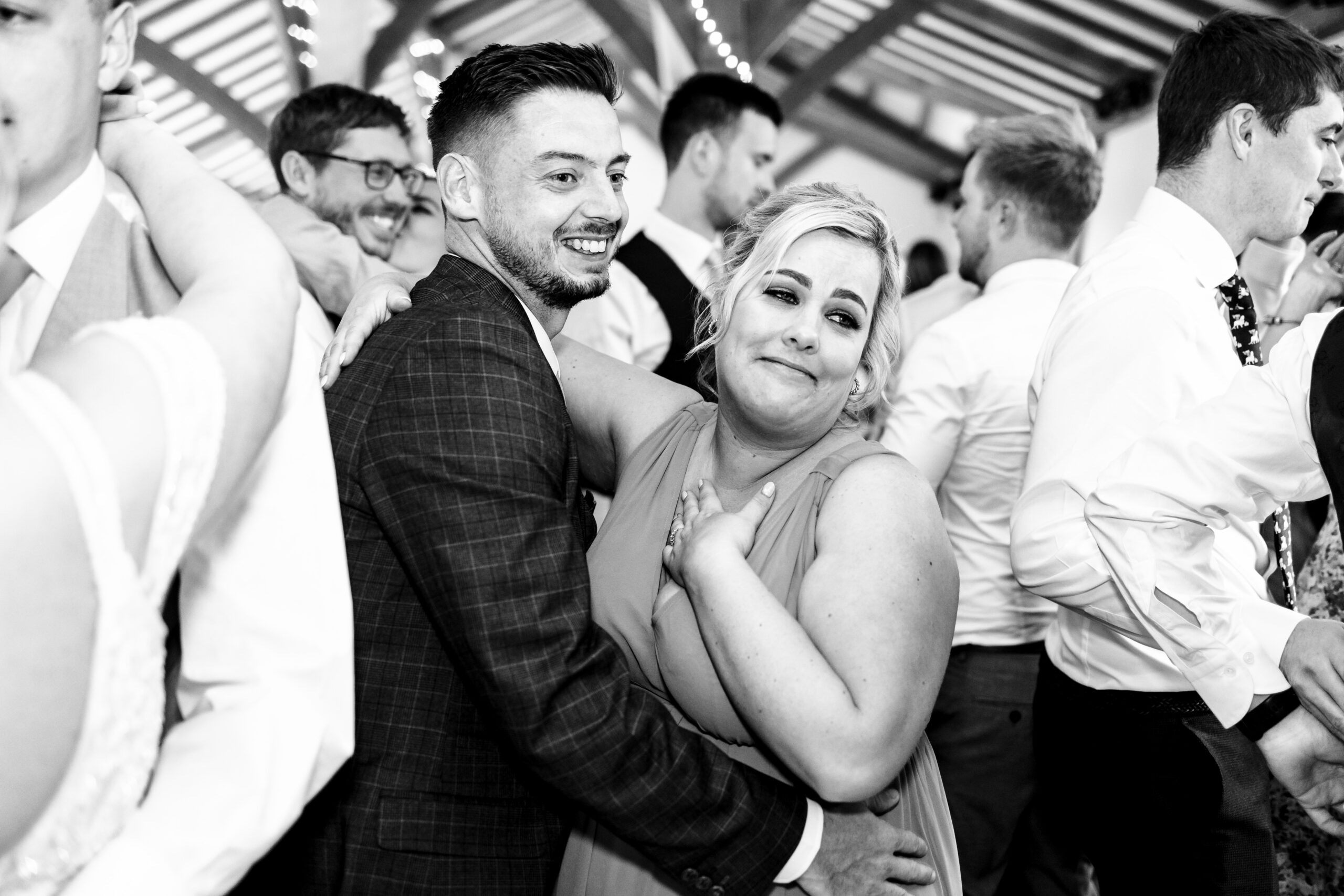 A black and white photo of two guests in an embrace as they dance. They are smiling and the lady has her hand on her heart endearingly. 