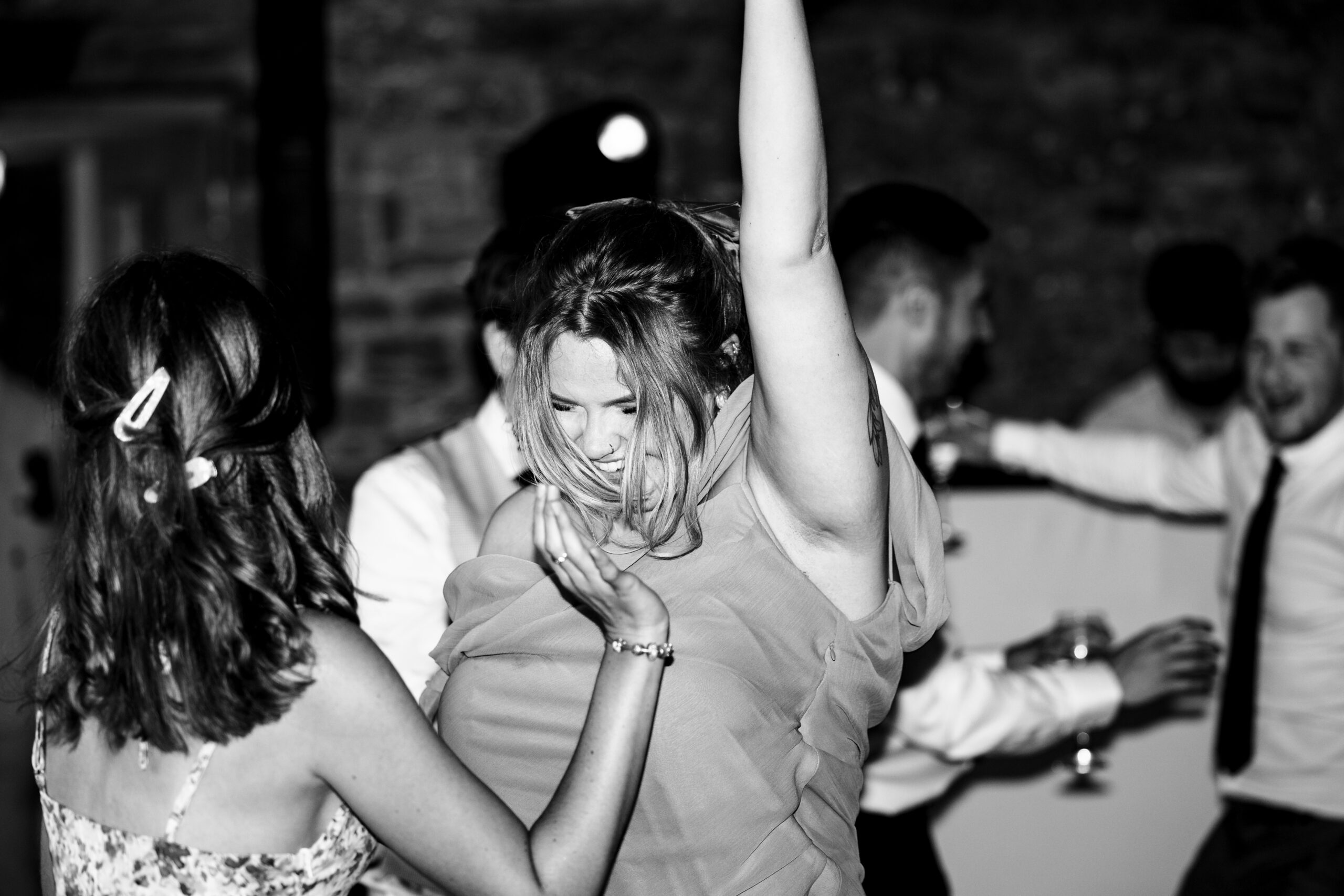 A black and white photo of two wedding guests. One is smiling with her arm in the air as she dances the night away.