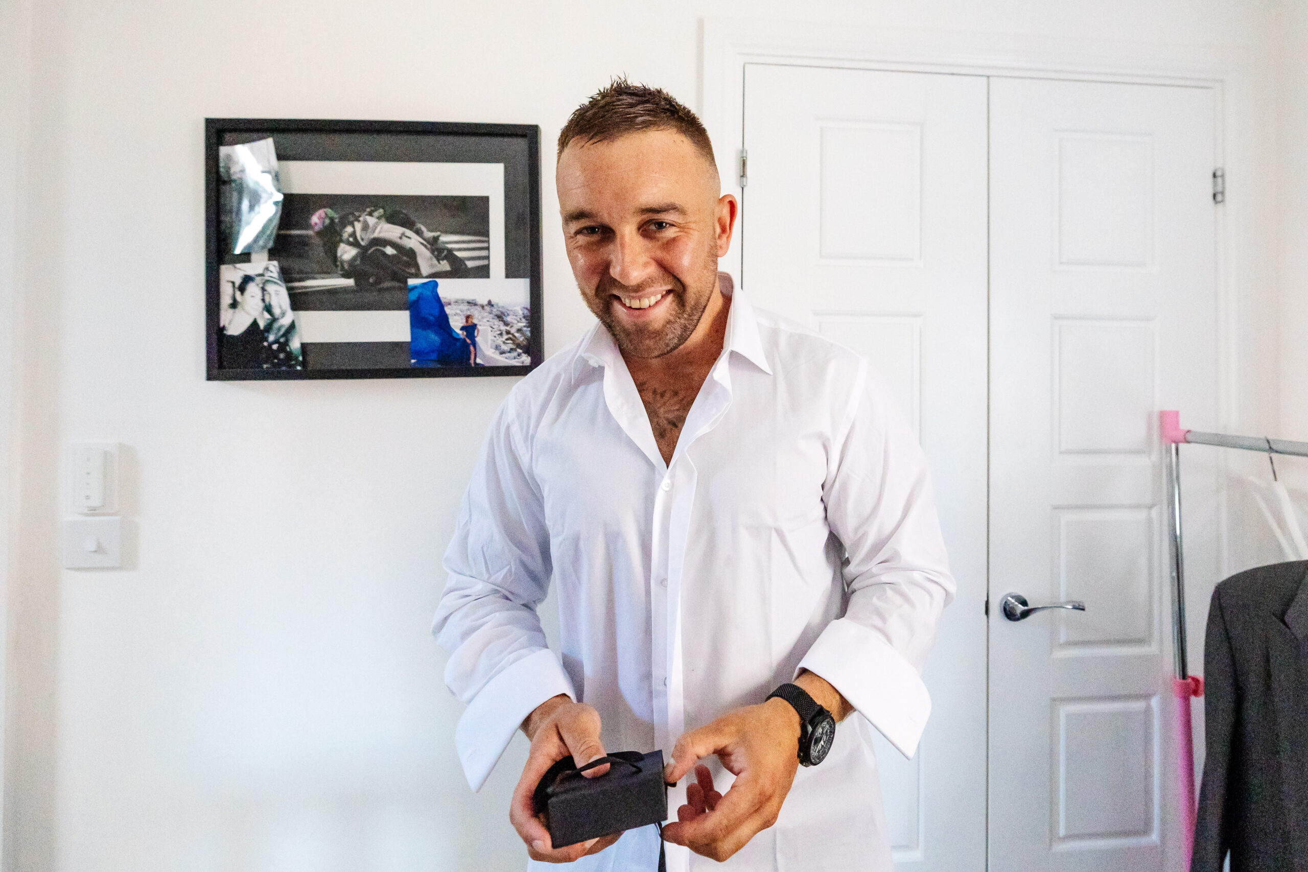 A groom in a white shirt holding a bxo in his hand. He has his top button undone and is wearing a black watch and is smiling.