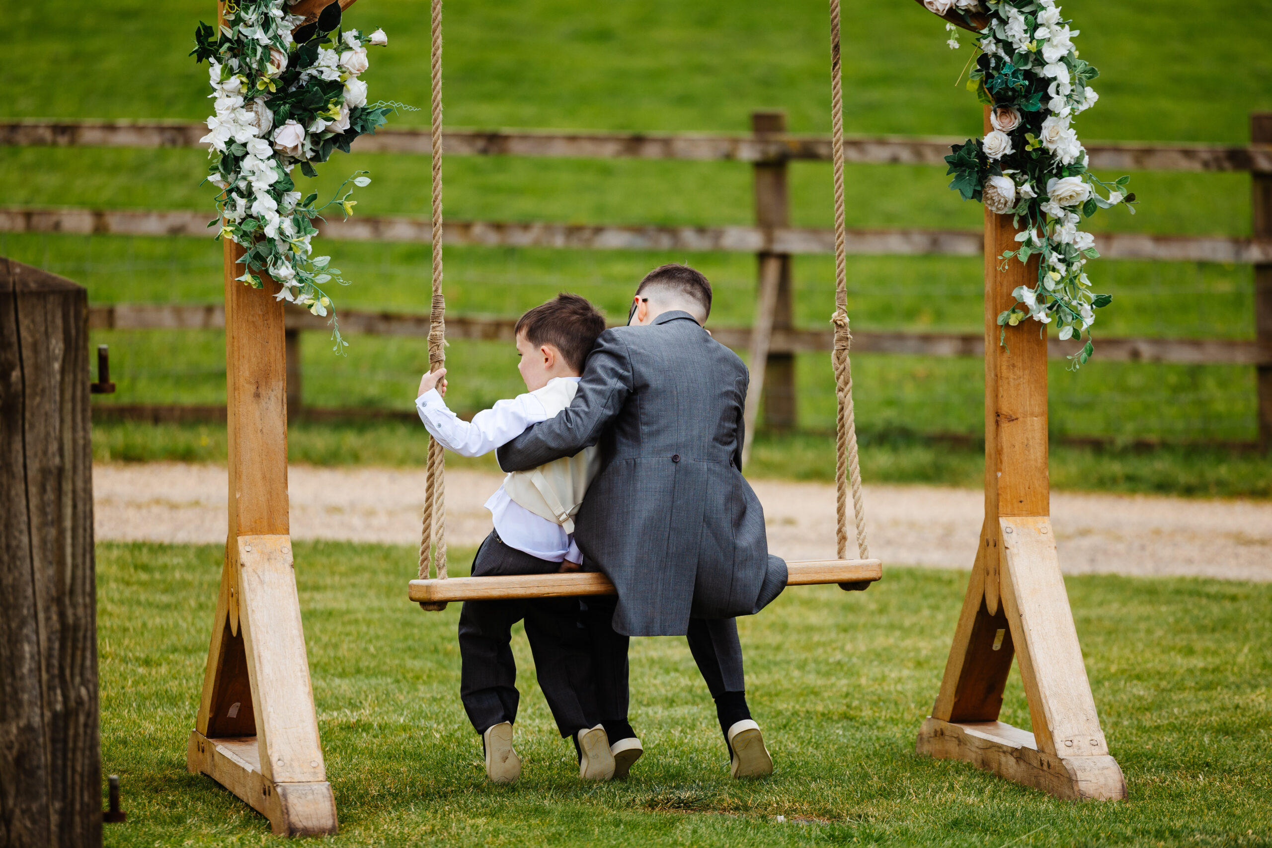 A photo of a wooden swing seat with white flowers as decoration. There are two boys sitting on it. The eldest boy has his arm around the smaller one and the smaller one has his hand on the side for stability.