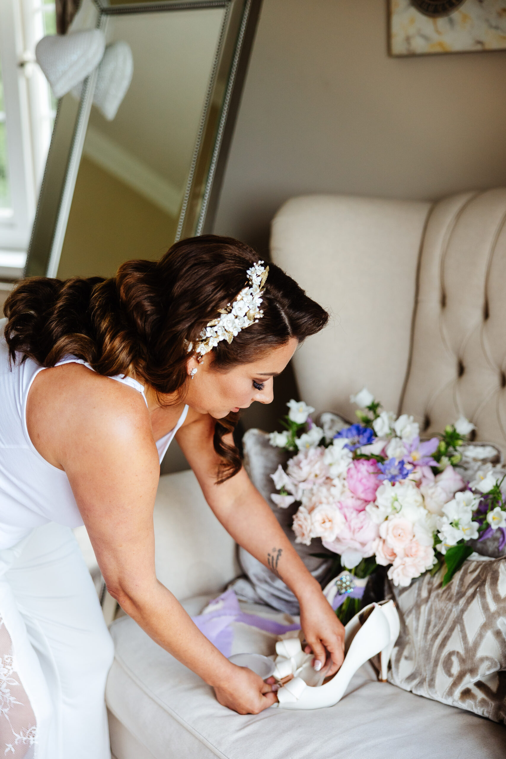 A bride in white pyjamas going to her wedding shoes. She is looking down at them, adjusting them.